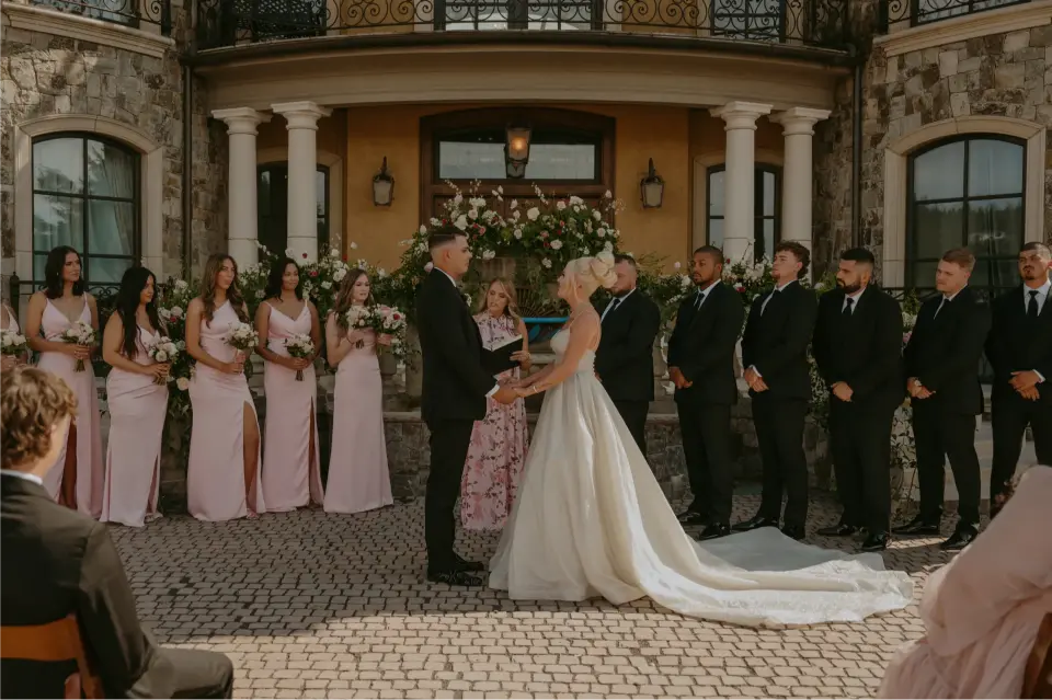 Bride and groom exchanging vows during an elegant outdoor Chateau de Harpe wedding ceremony with their wedding party standing beside them.