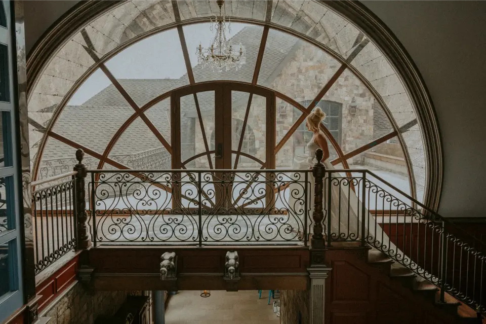 Bride standing by the grand arched window and elegant staircase inside Chateau de Harpe during a luxury wedding.