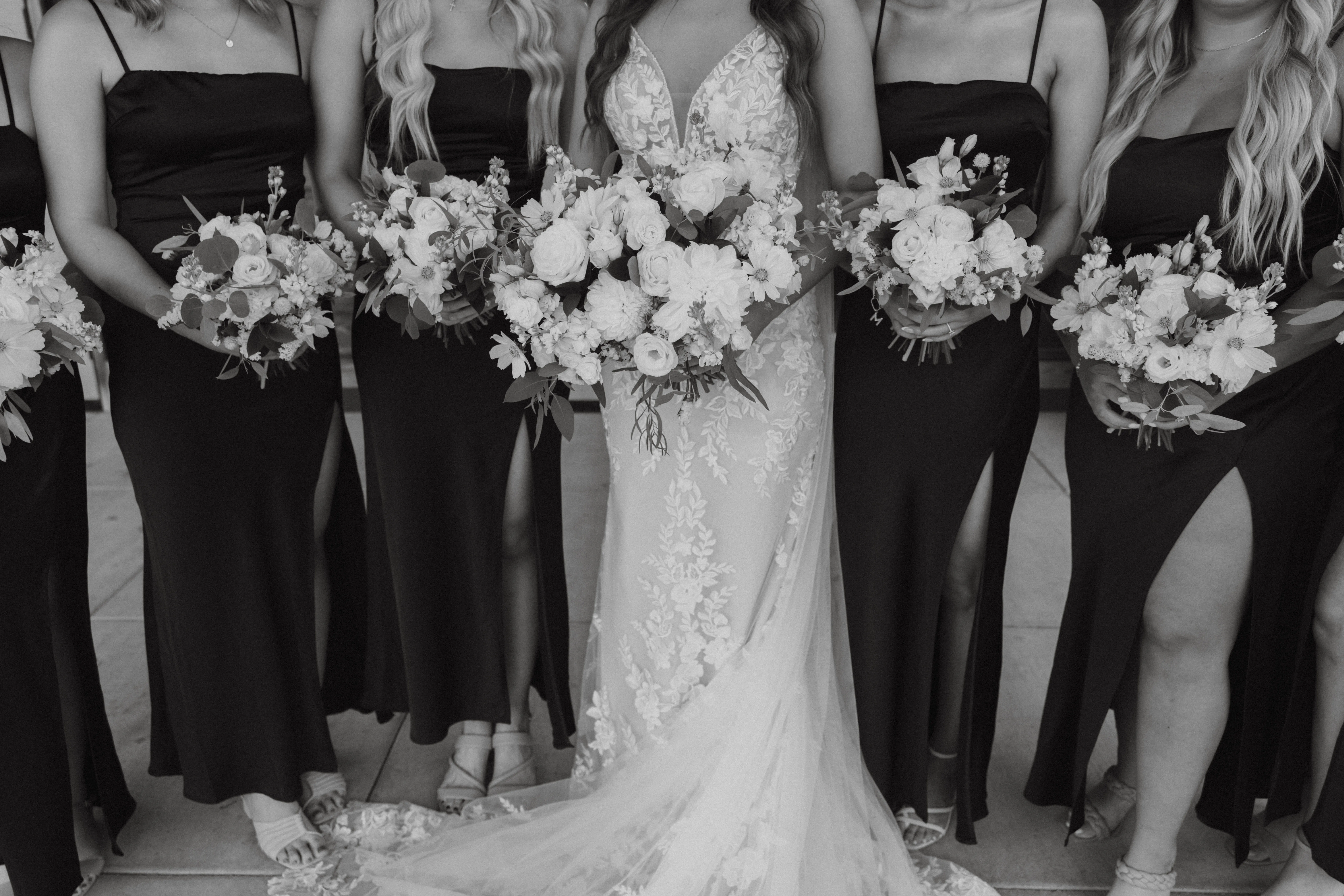 Bride in a lace wedding gown standing with bridesmaids in black dresses, each holding white floral bouquets during an elegant wedding portrait.