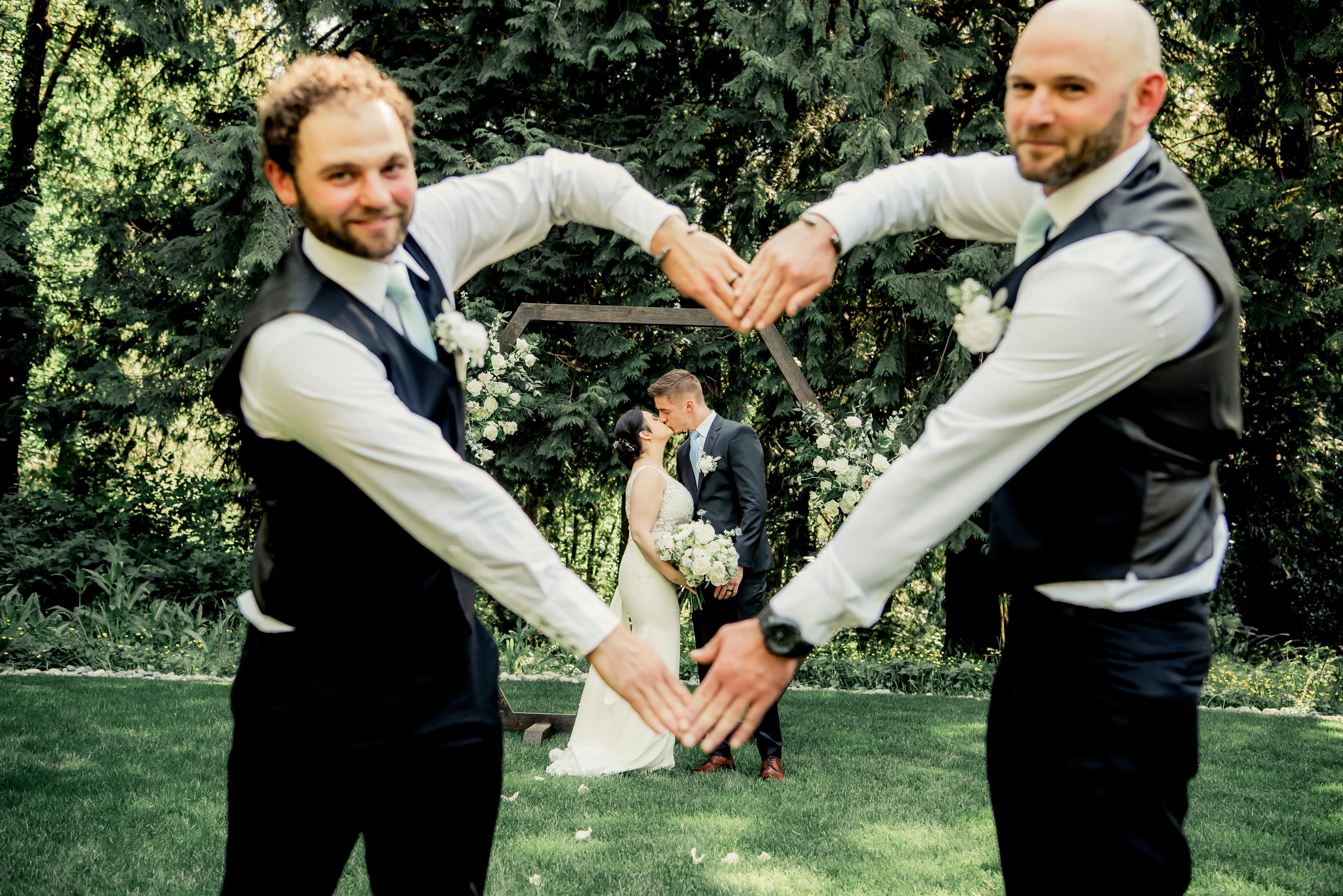 Two groomsmen form a heart shape with their arms, framing the bride and groom sharing a kiss beneath a floral wedding arch during an outdoor wedding ceremony.