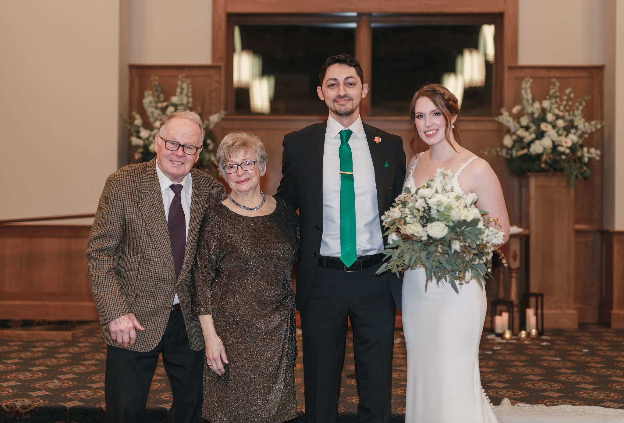 Bride and groom posing with elderly family members during a formal indoor wedding portrait, with the bride holding a white and greenery bouquet in a warmly decorated ceremony venue