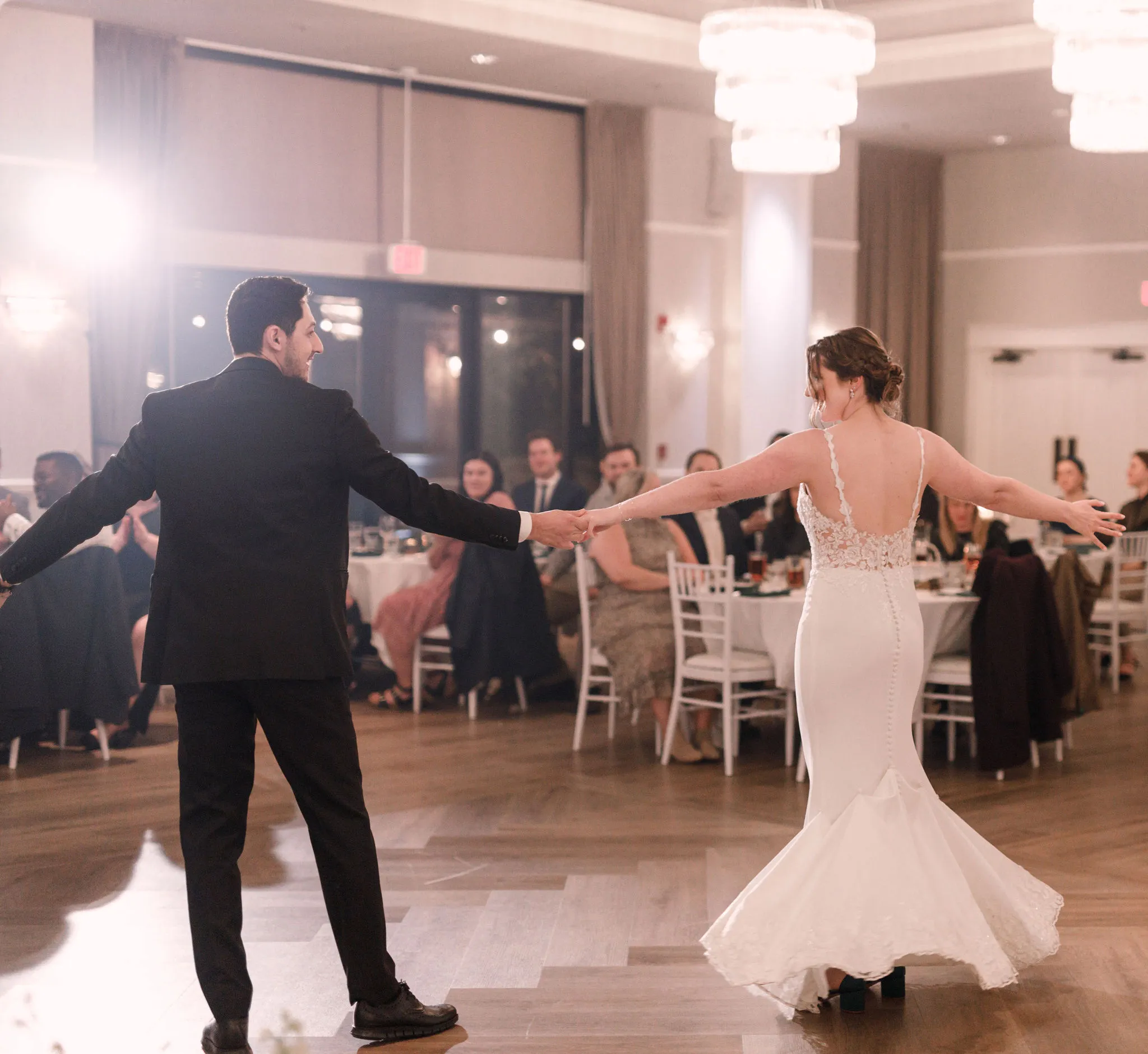 Bride and groom holding hands during their first dance at an elegant indoor wedding reception, surrounded by guests and warm chandelier lighting.