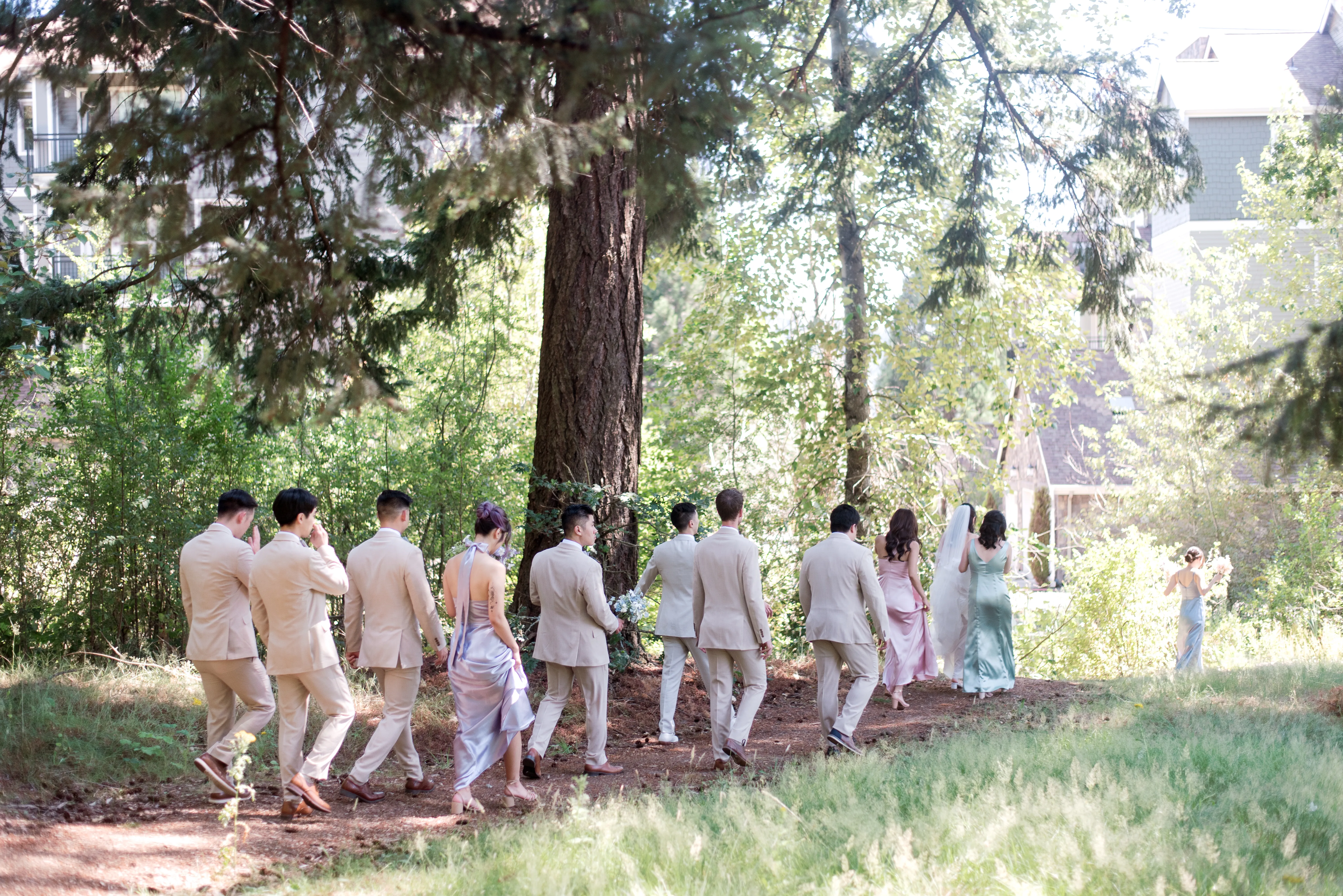 Bride and groom walking hand in hand through a forest with their wedding party, featuring pastel bridesmaid dresses, beige suits, and colorful floral bouquets during an outdoor wedding celebration.