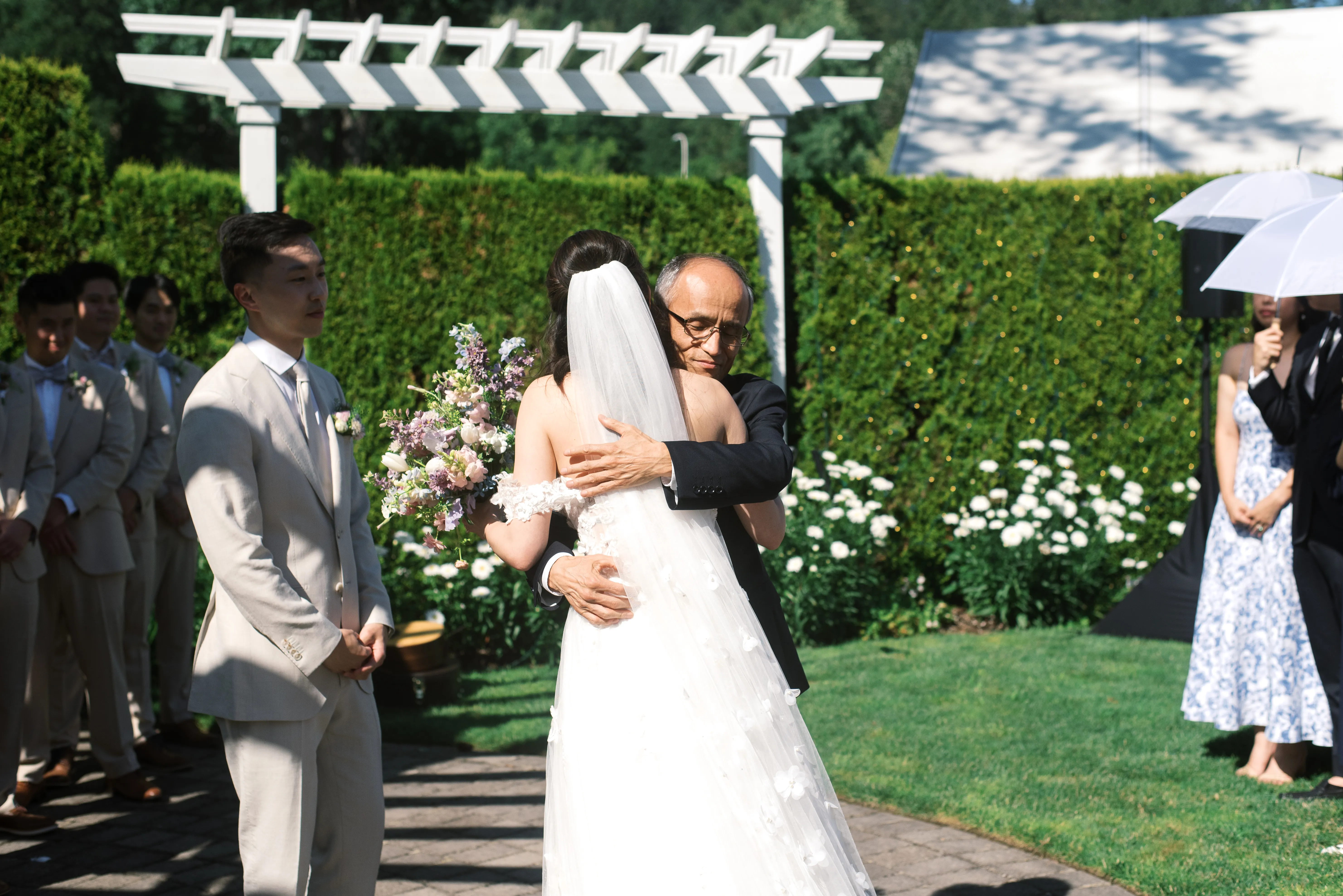 Bride embracing her father during an emotional outdoor wedding ceremony while the groom and wedding guests look on beneath a white