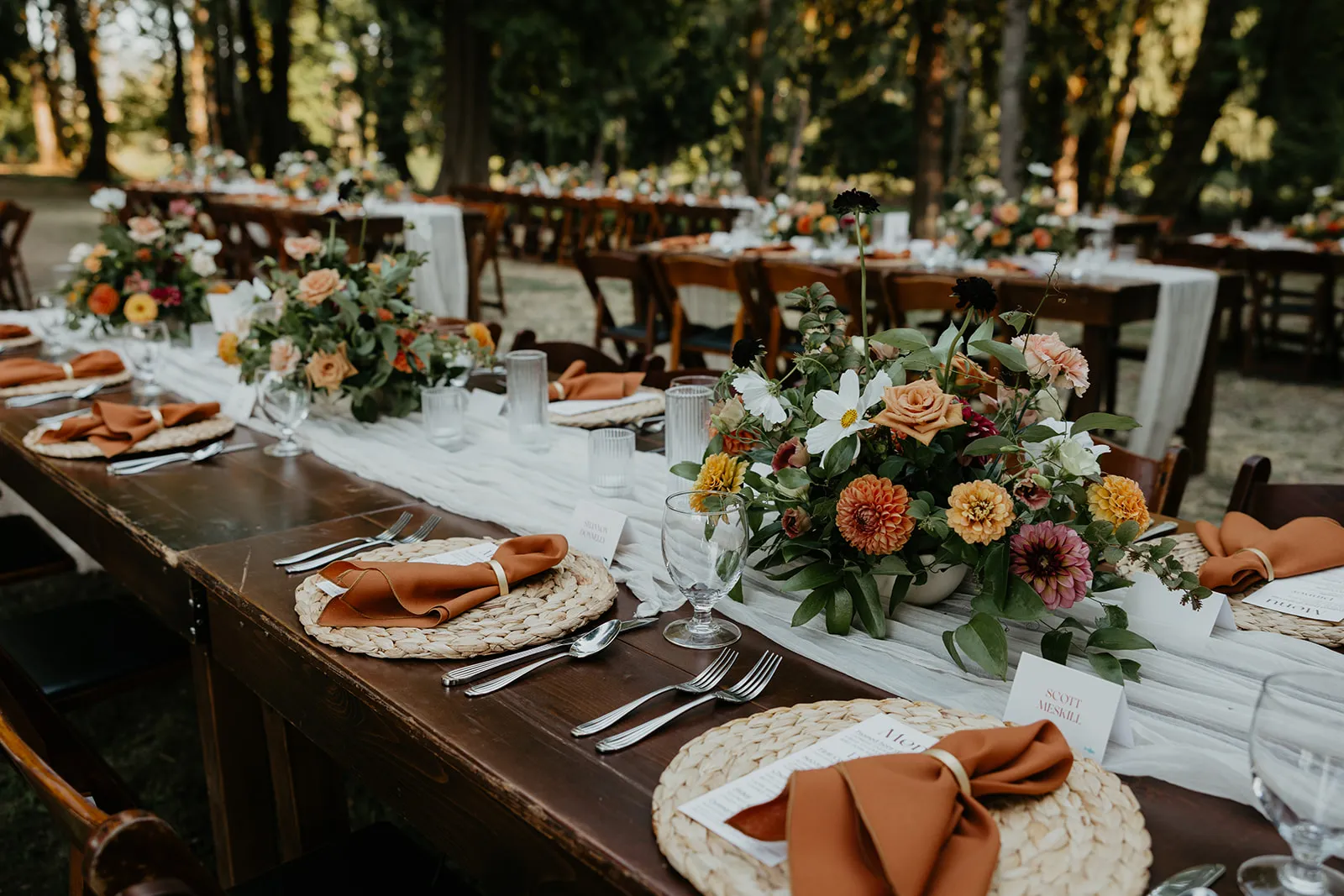 Elegant outdoor wedding reception tables with colorful floral centerpieces, wooden tables, rust-colored napkins, and place settings arranged beneath towering trees.