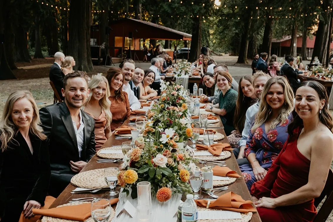 Wedding guests seated at long family-style reception tables with colorful floral centerpieces and elegant place settings during an outdoor woodland wedding reception.