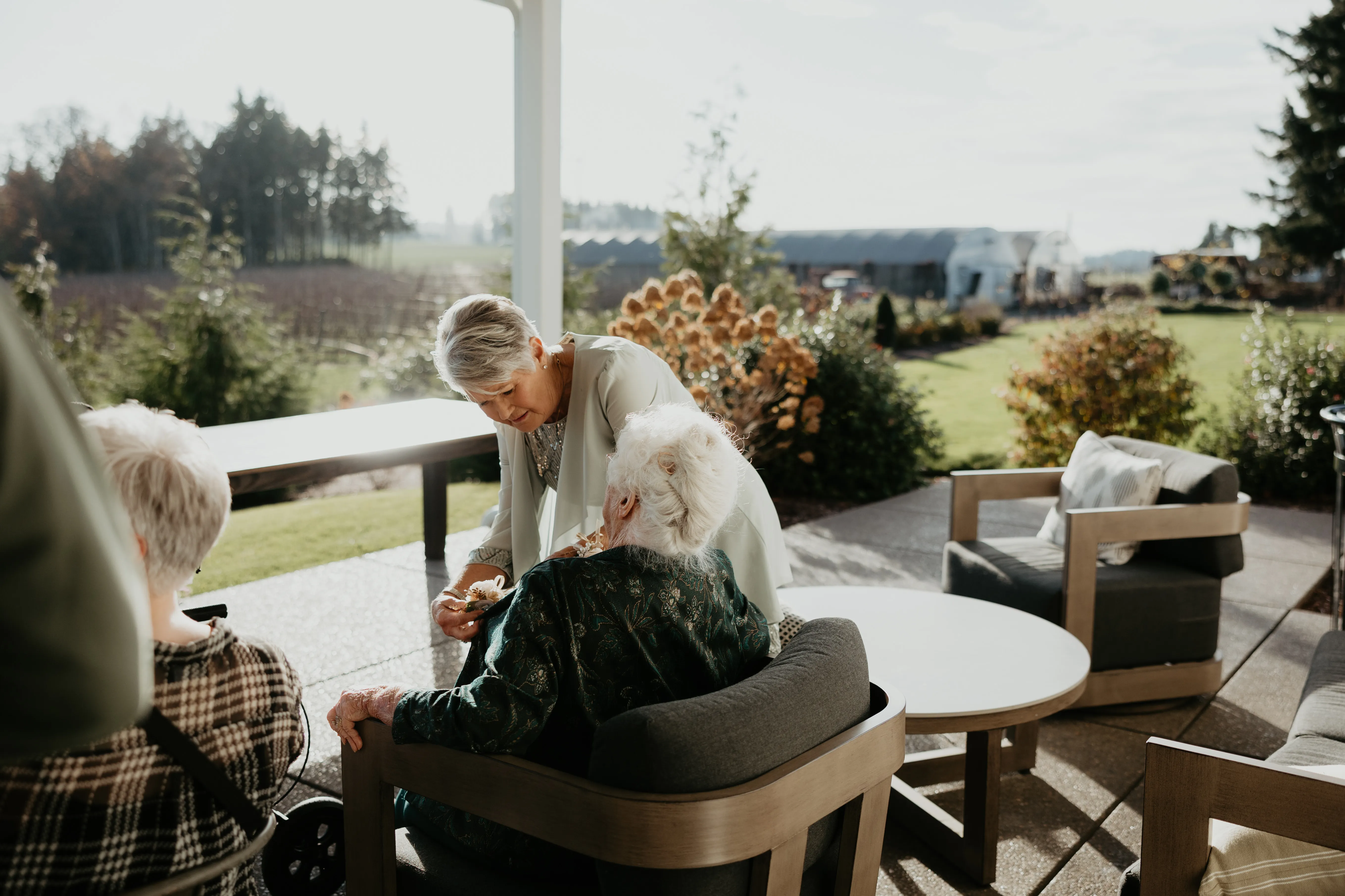 Family members sharing a heartfelt moment on an outdoor patio overlooking gardens and vineyards before a wedding celebration, with elderly guests seated in comfortable lounge chairs.