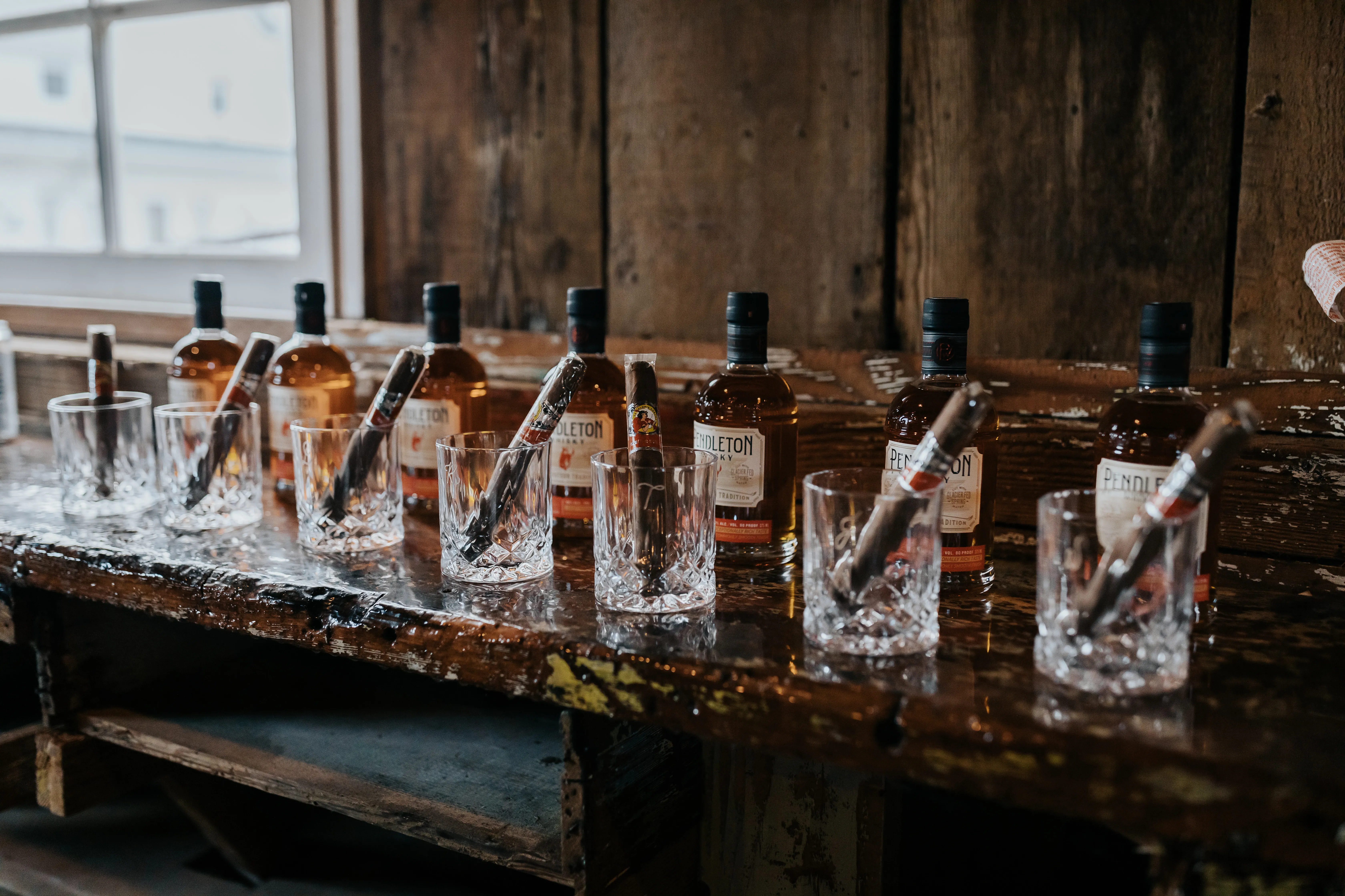 Rustic wedding whiskey and cigar station featuring Pendleton whiskey bottles, crystal whiskey glasses, and premium cigars displayed on a wooden bar for guests.