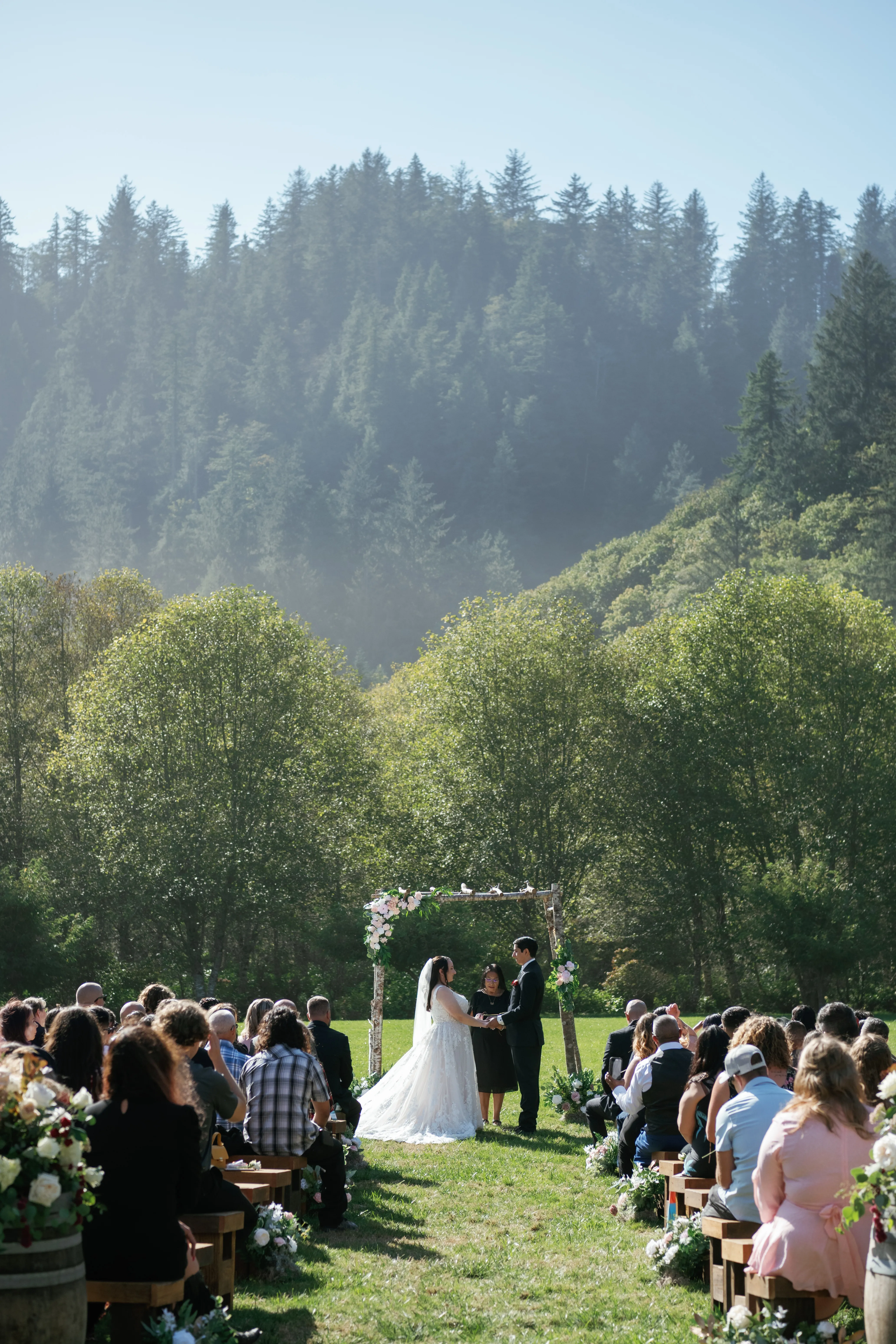 Bride and groom exchanging vows beneath a floral wedding arch during an outdoor ceremony, surrounded by guests with a scenic forest and mountain backdrop.