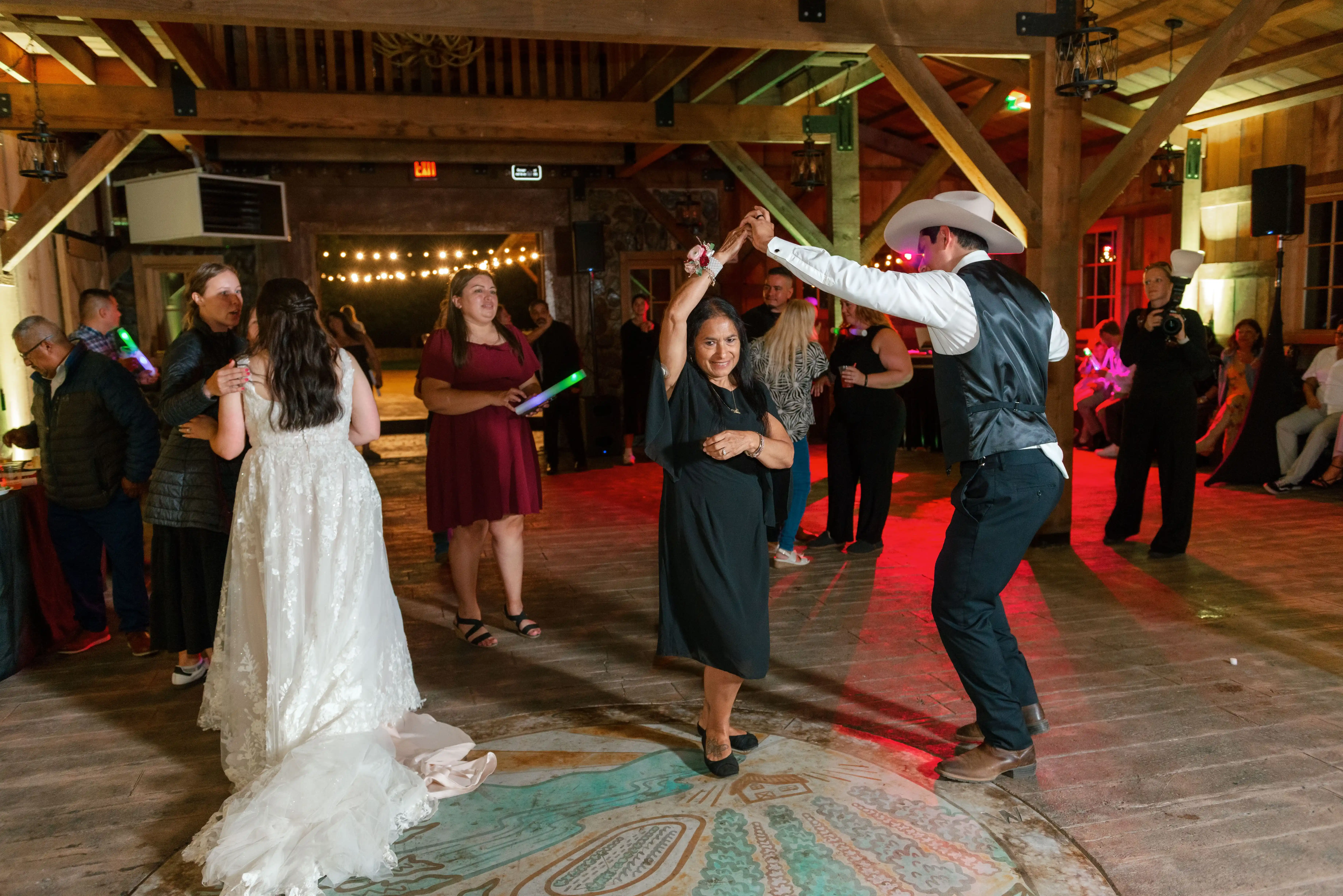 Wedding guests dancing during a lively rustic barn reception, with a guest in a cowboy hat twirling a family member on the dance floor under warm string lights.
