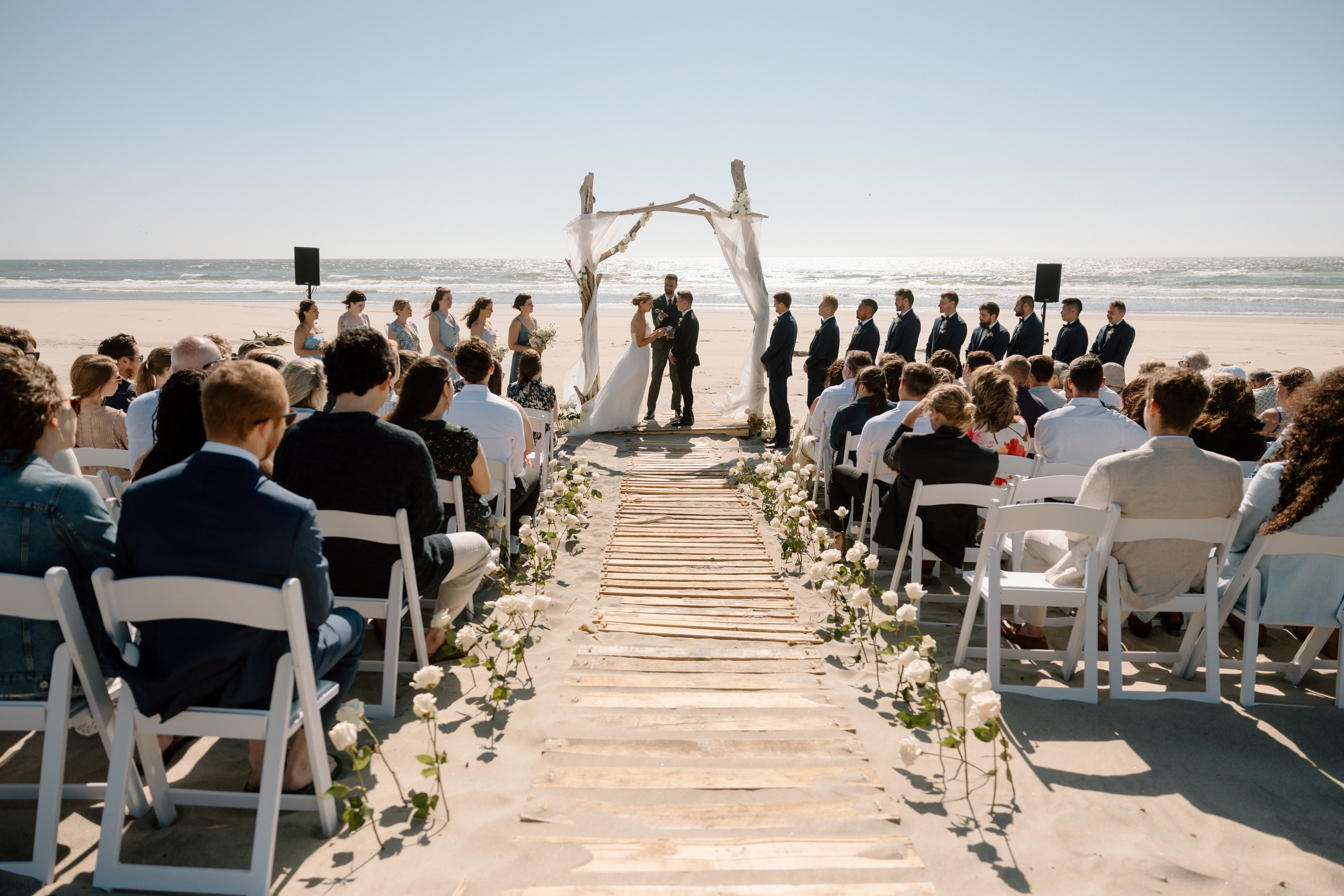 Guests seated for a beach wedding ceremony as the bride and groom exchange vows beneath a driftwood arch with ocean waves and a clear blue sky in the background.