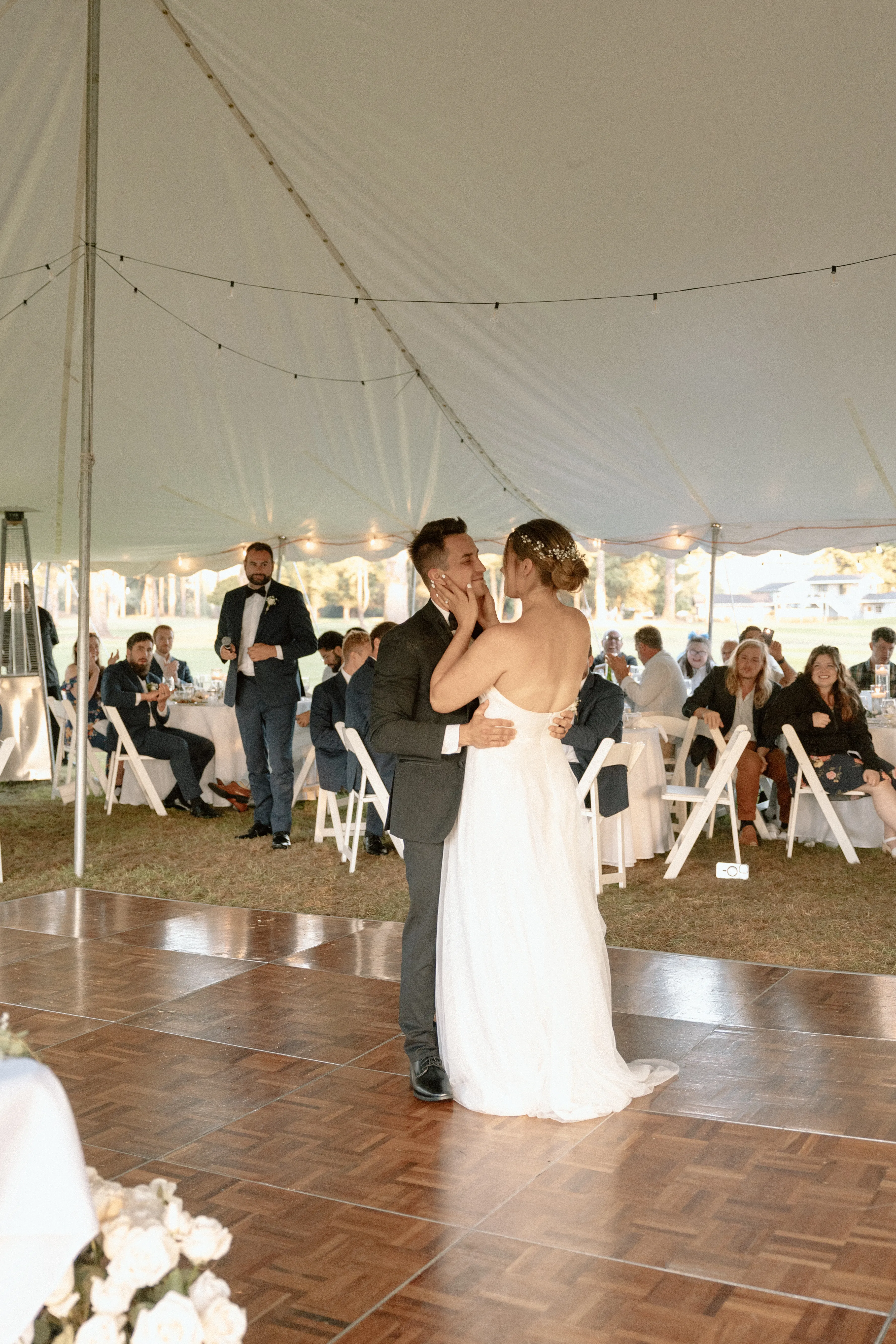 Guests seated for a beach wedding ceremony as the bride and groom exchange vows beneath a driftwood arch with ocean waves and a clear blue sky in the background
