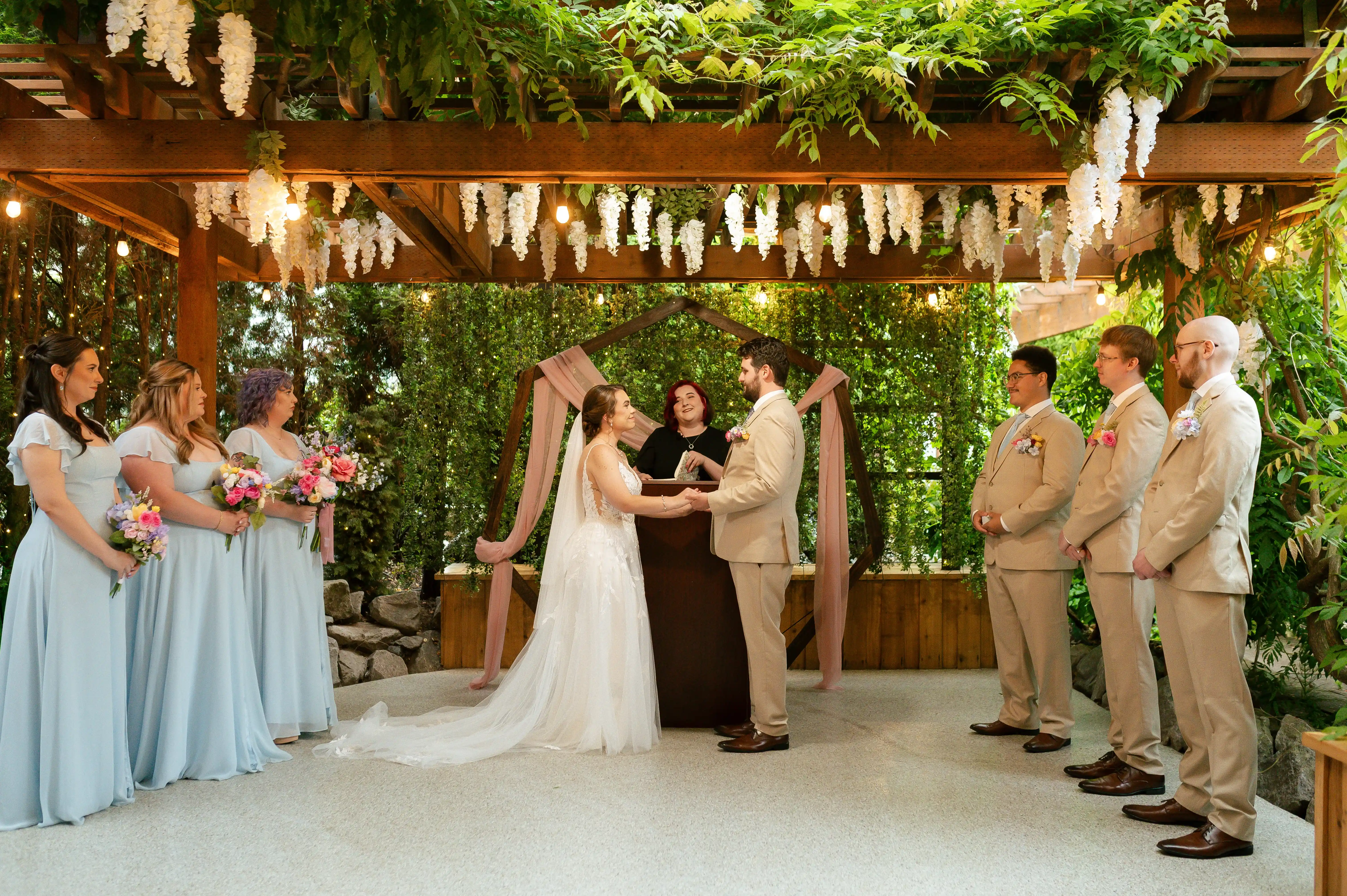 Bride and groom exchanging vows beneath a geometric floral arch under a flower-covered pergola, surrounded by bridesmaids and groomsmen during an elegant garden wedding ceremony.