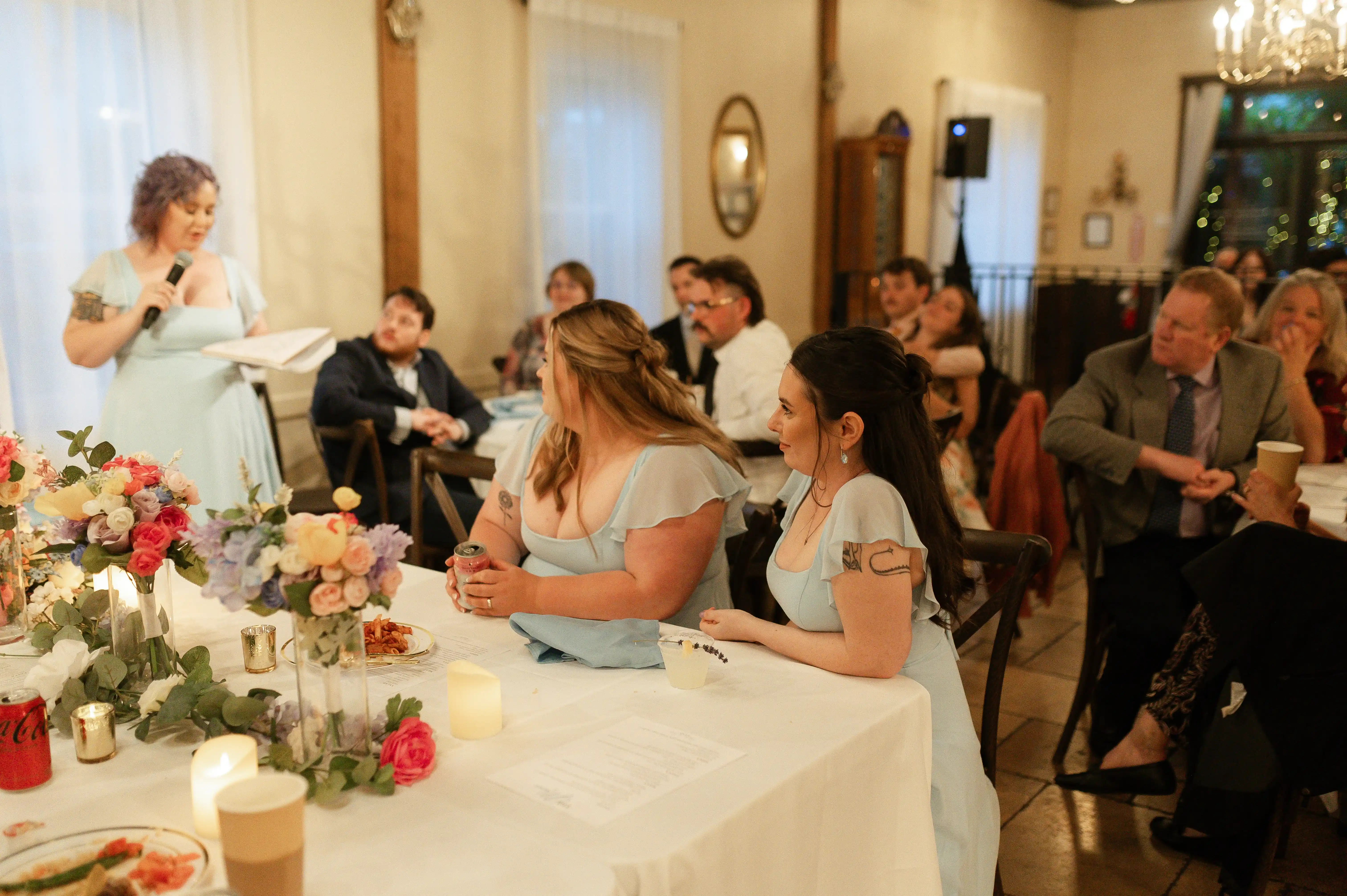 Bridesmaid giving a heartfelt wedding reception speech while guests and the bridal party listen around elegantly decorated tables with colorful floral centerpieces.