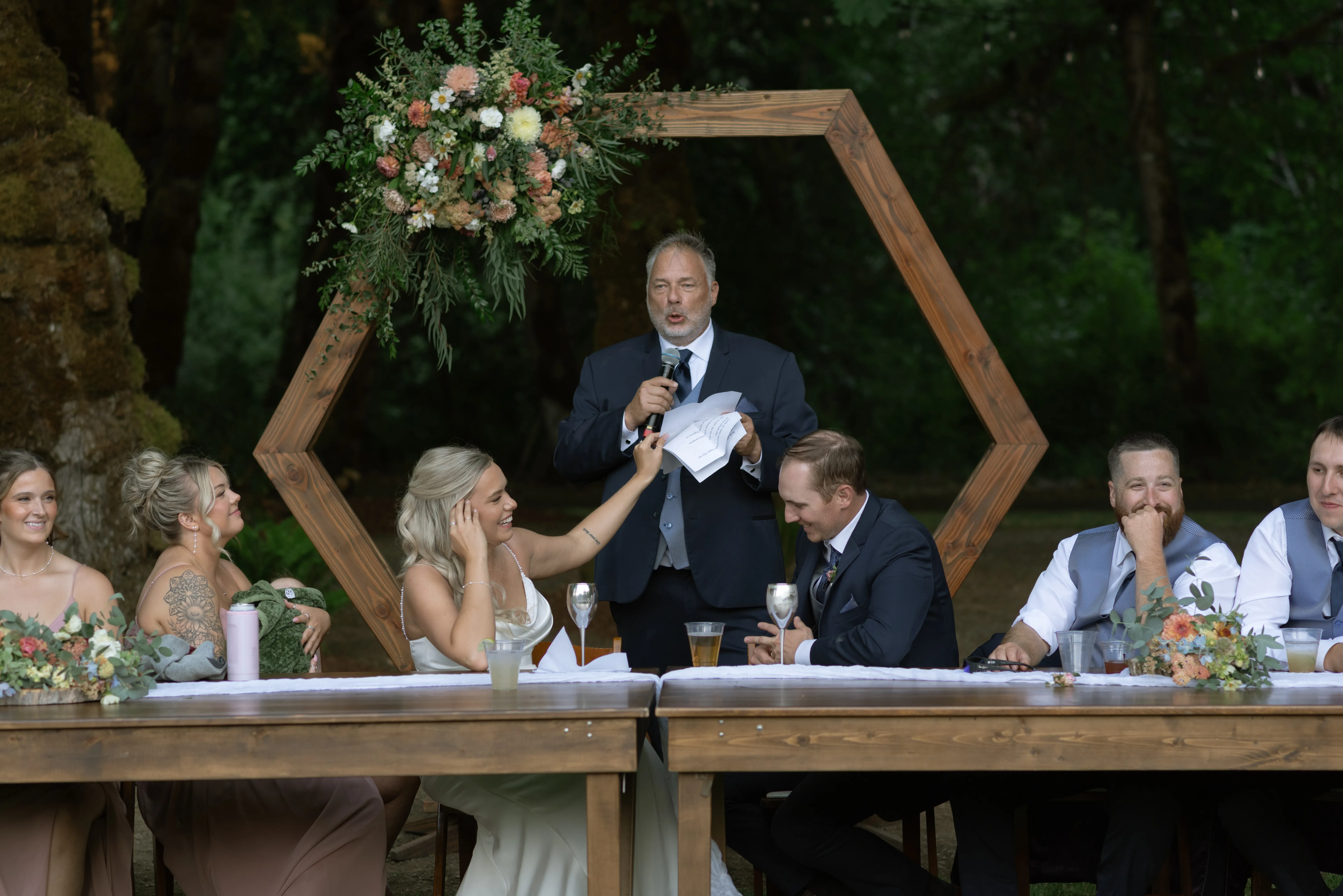Father of the bride giving a heartfelt wedding toast while the bride, groom, bridal party, and guests smile beneath a geometric floral arch during an outdoor wedding reception.