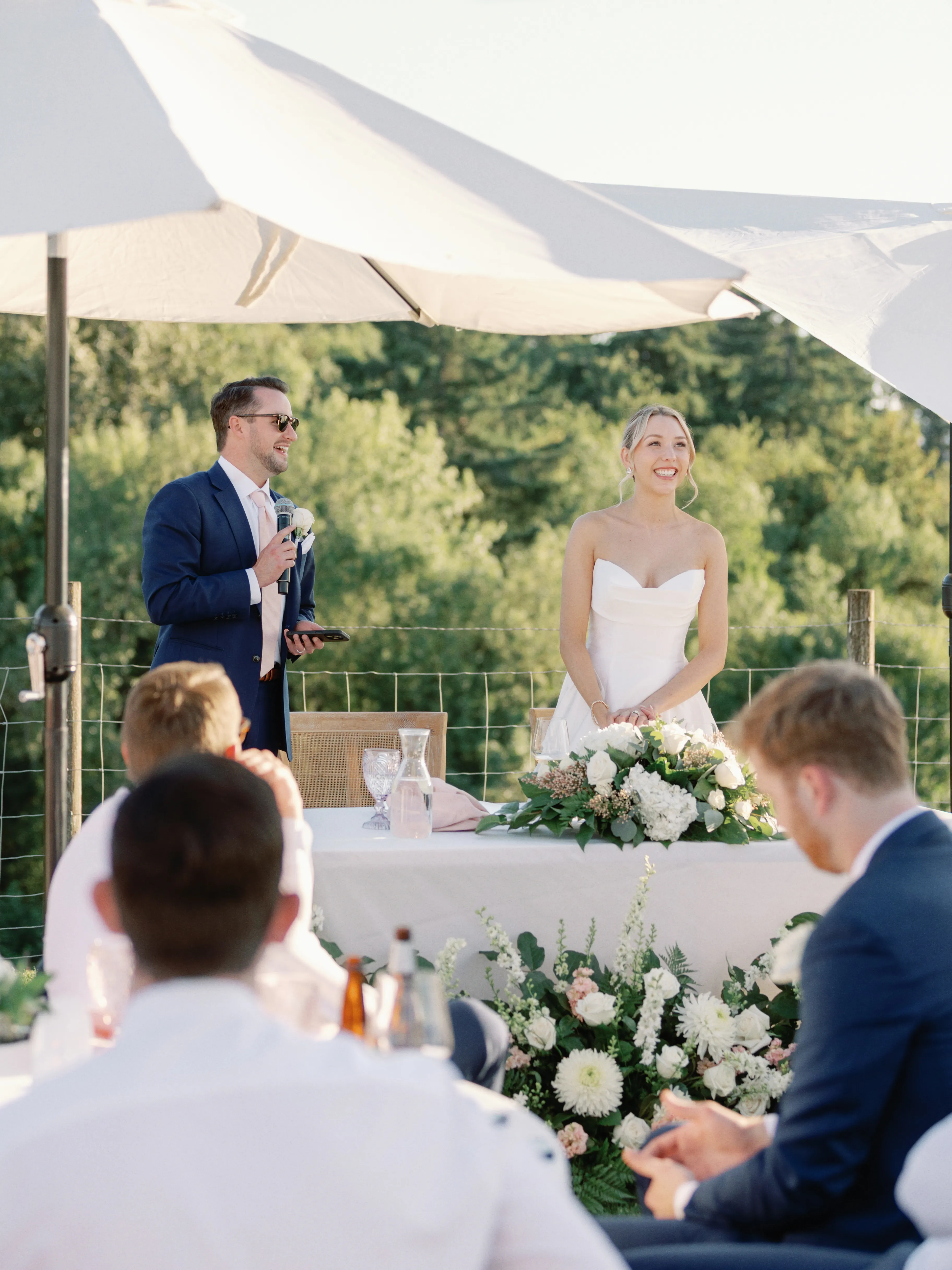 Bride smiling during an outdoor wedding reception while a guest gives a heartfelt toast, with elegant white floral arrangements and guests gathered beneath sun umbrellas.