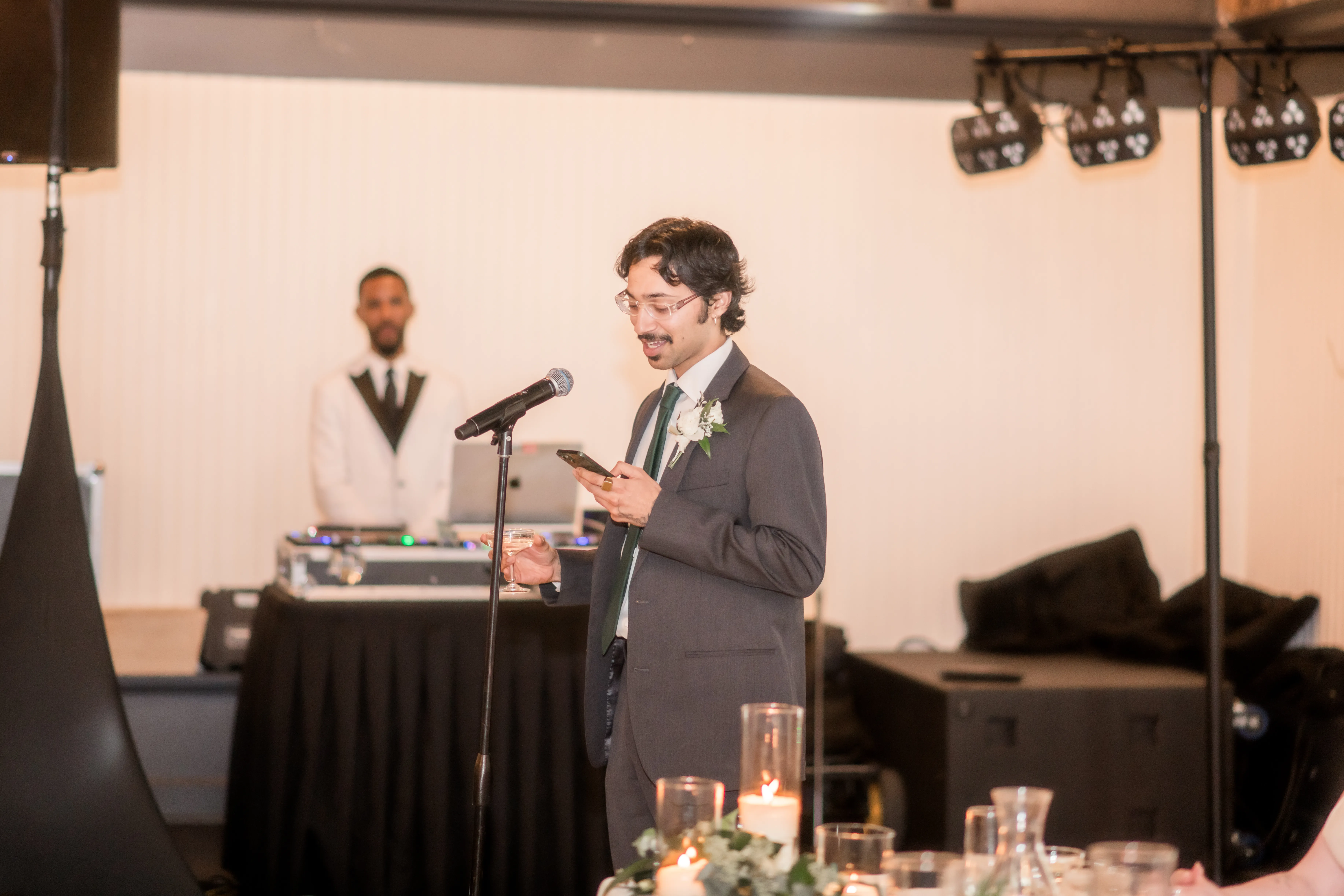 Best man giving a heartfelt wedding reception speech while holding a champagne glass, reading from his phone, with the DJ setup in the background and guests listening during the celebration.