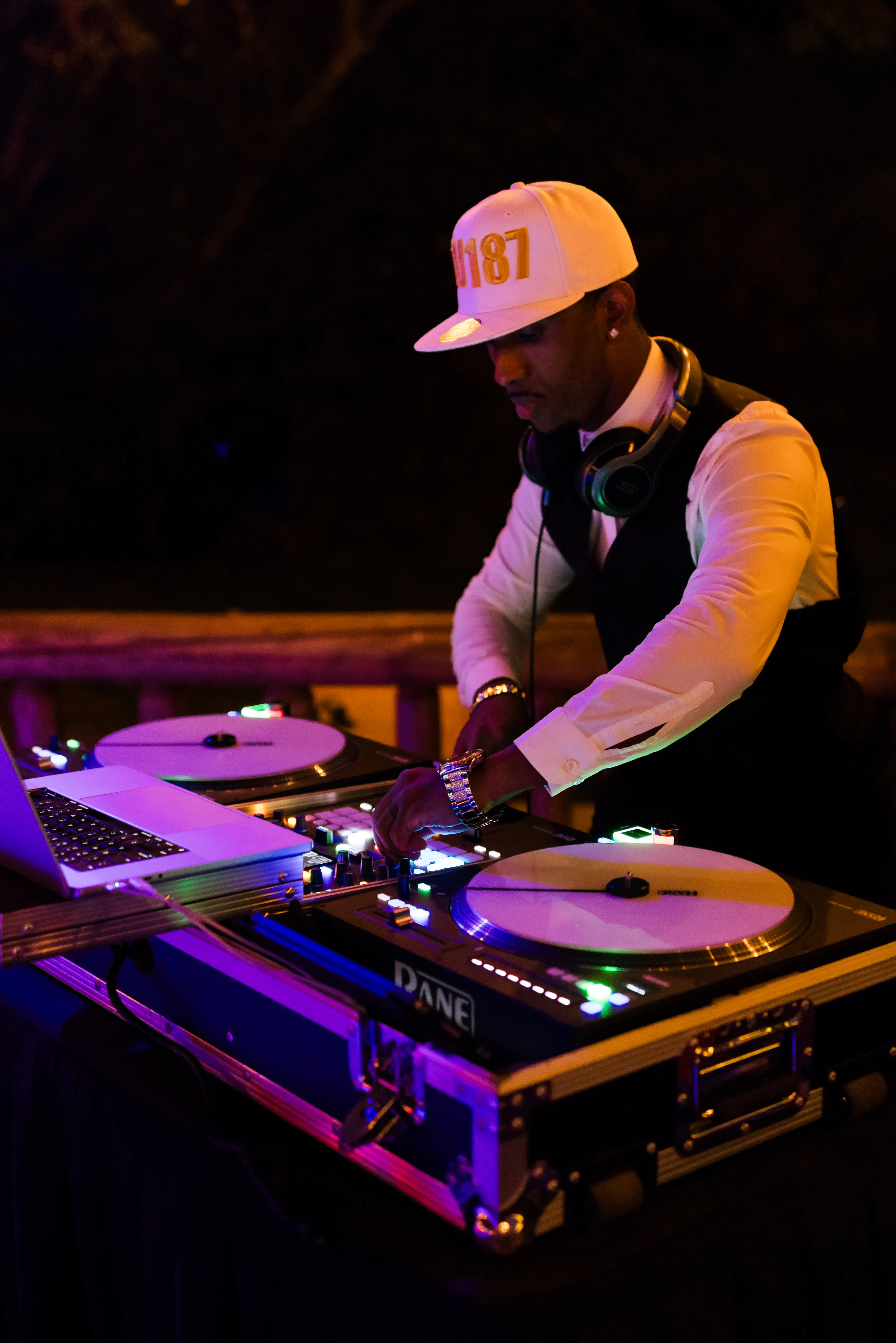 Professional wedding DJ mixing music on turntables and a laptop during an evening wedding reception, creating an energetic dance floor atmosphere for guests.