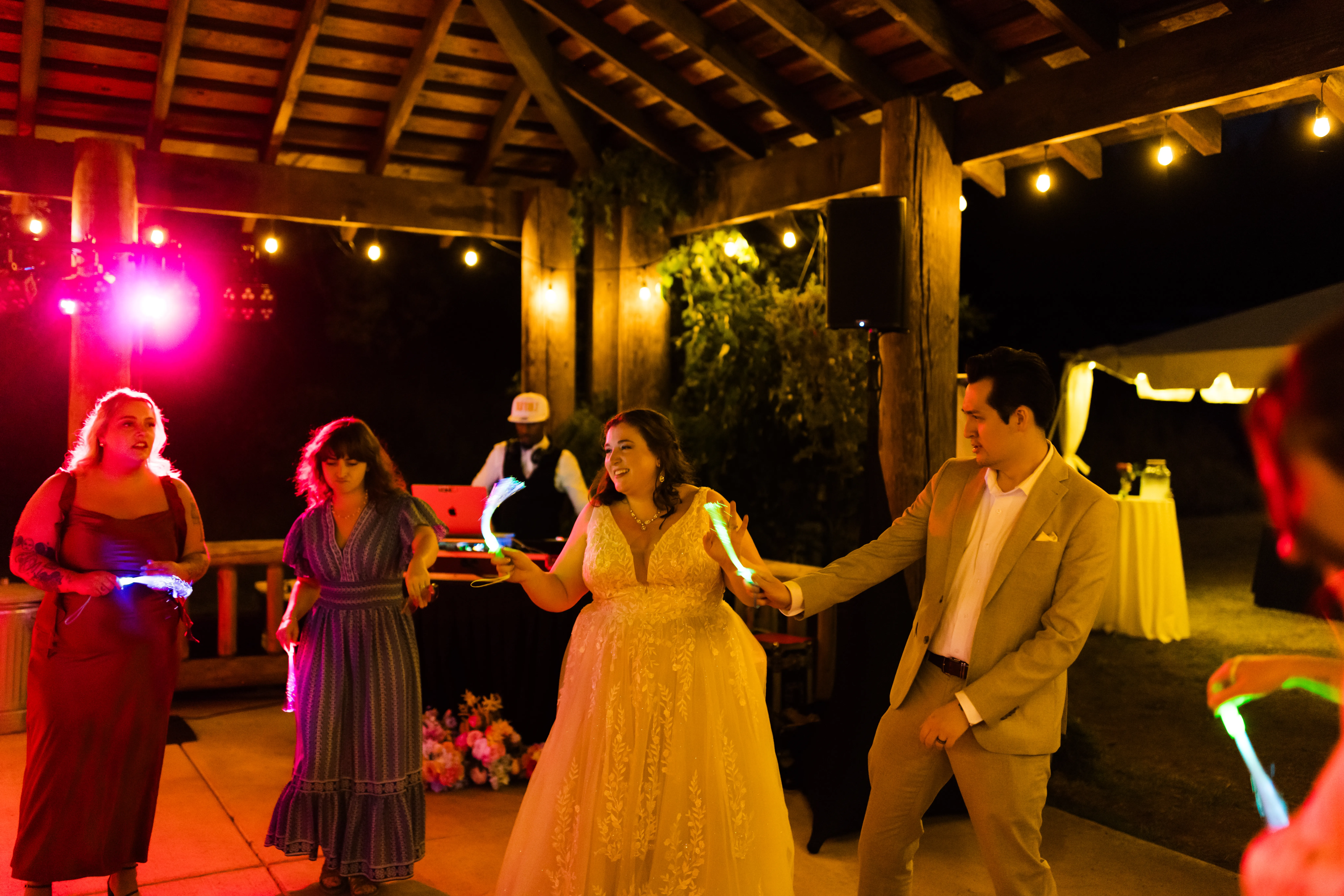 Bride and groom dancing with guests holding glow sticks during a lively wedding reception, while a professional DJ entertains under string lights at an outdoor venue.