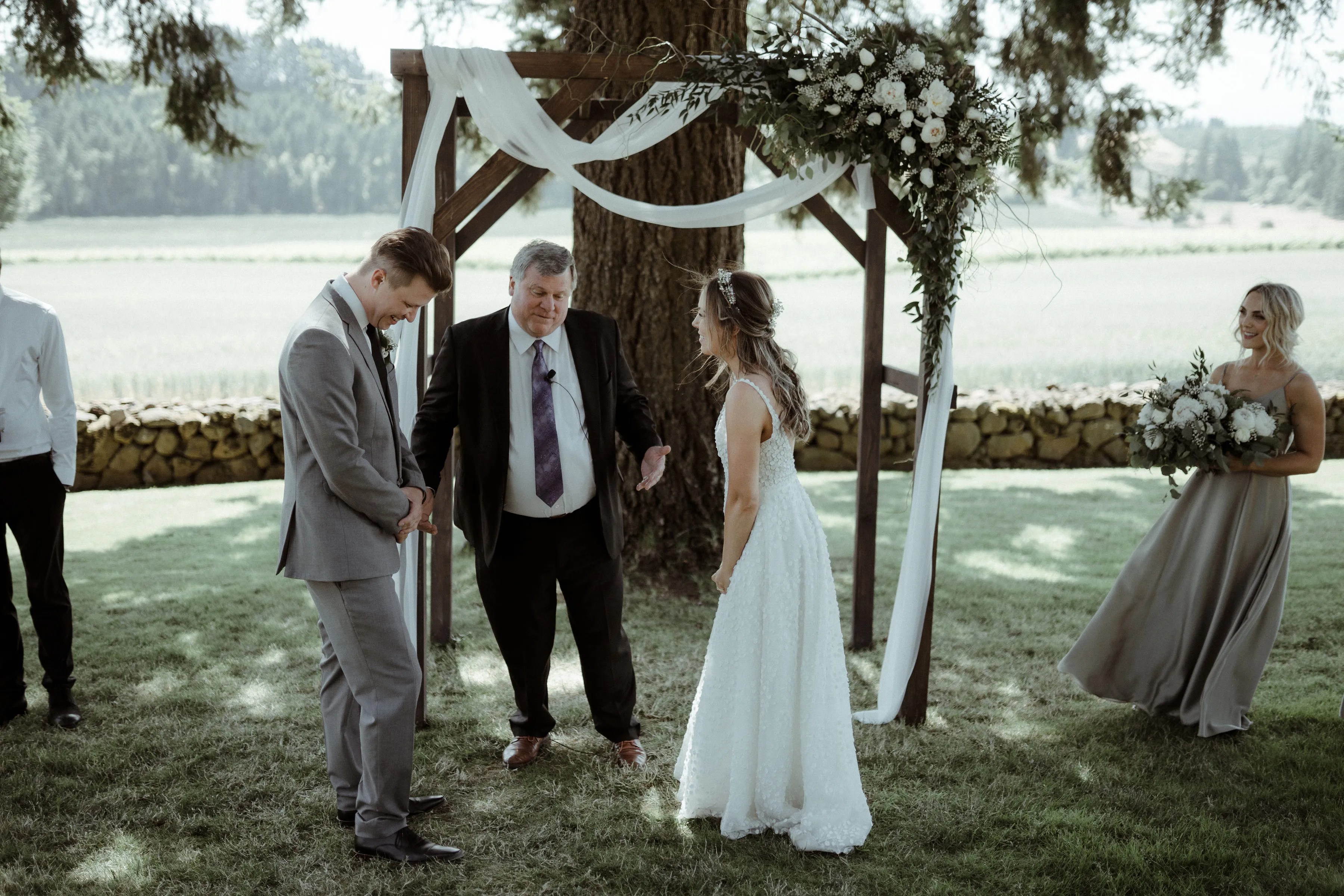 Bride and groom exchanging vows during a romantic outdoor lakeside wedding ceremony beneath a floral arbor, surrounded by their officiant and wedding party.