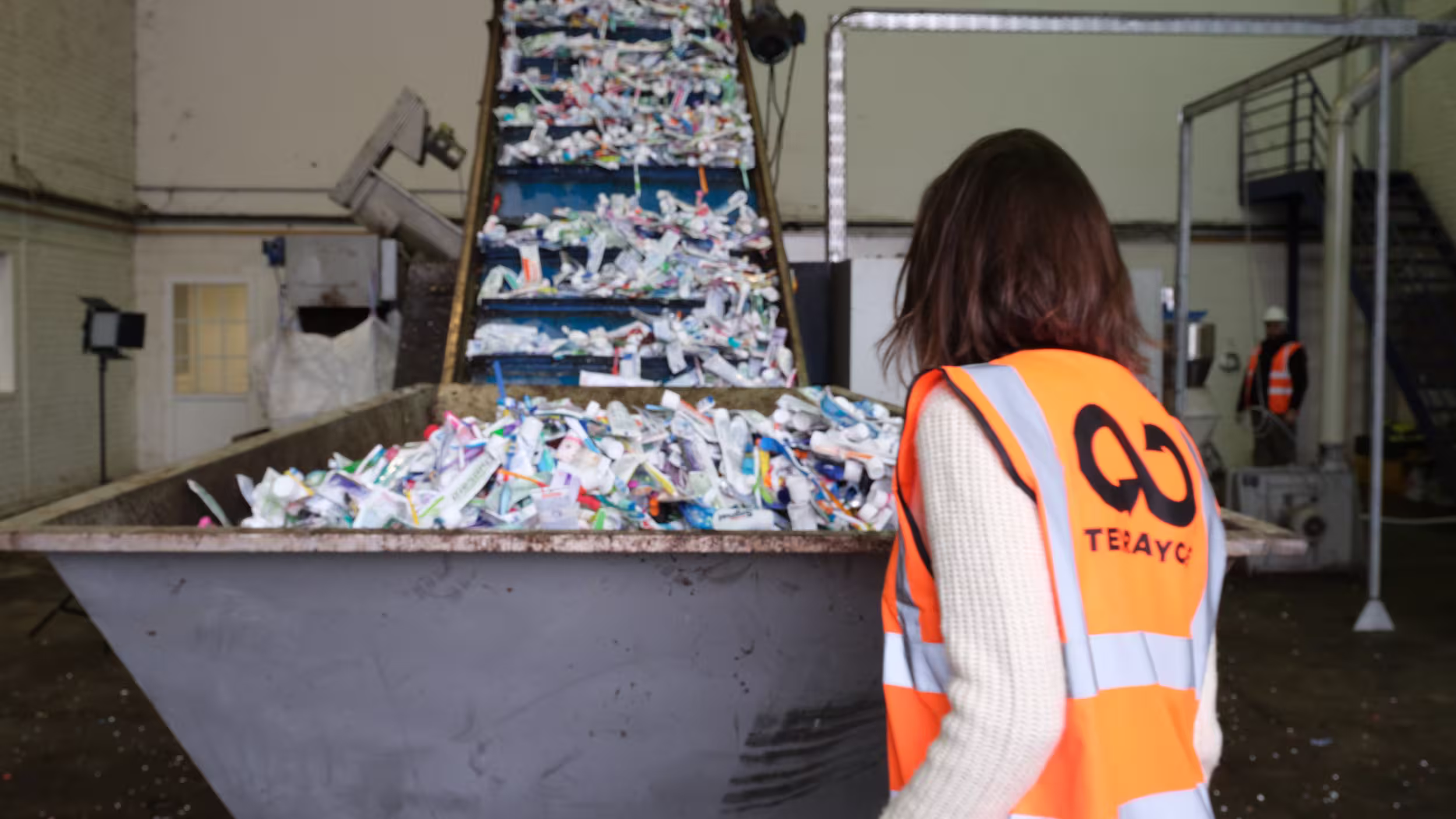 A woman in a TerraCycle safety vest observing a conveyor belt filled with used toothpaste tubes and hard-to-recycle plastics at a recycling facility