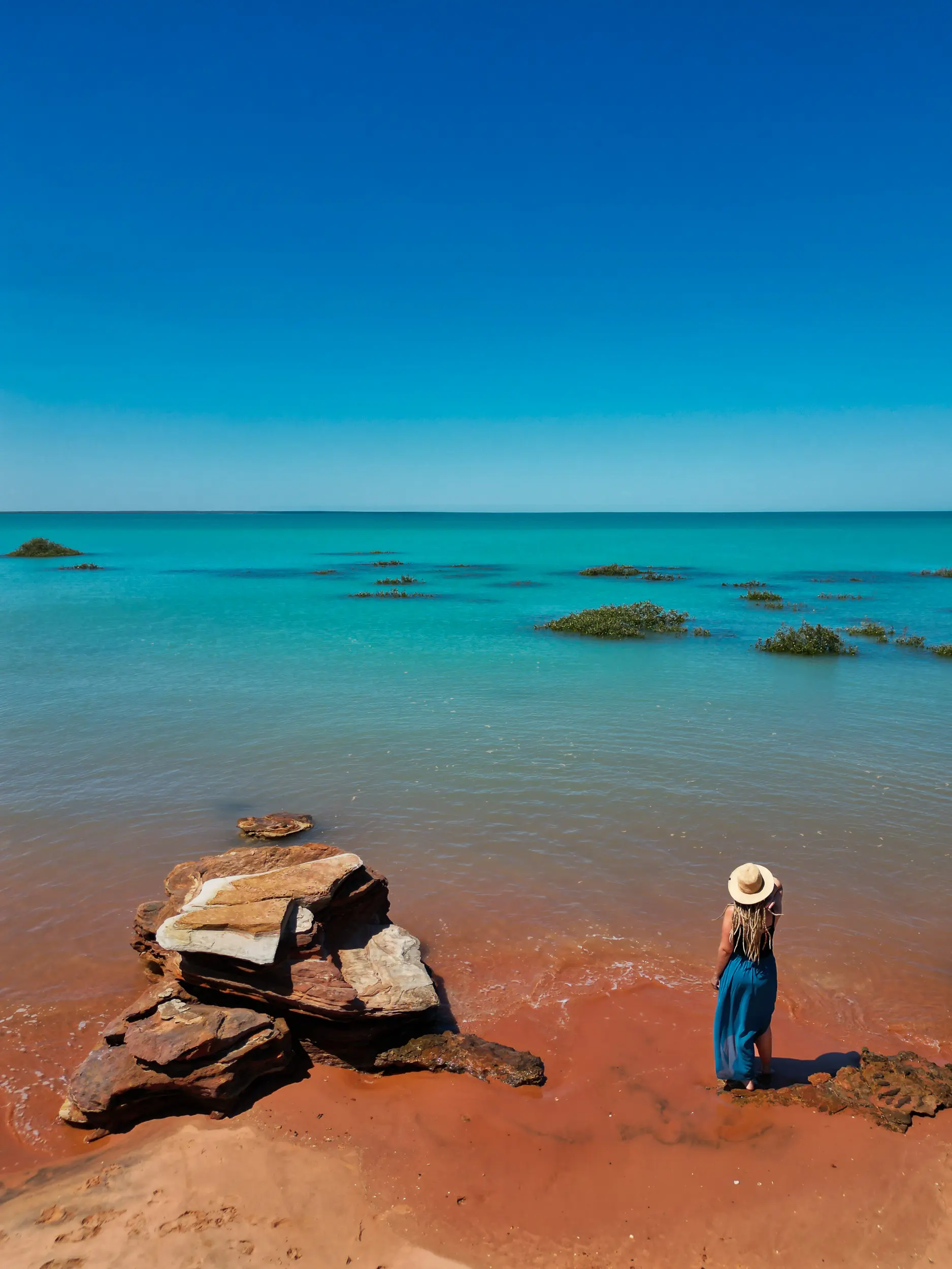 Woman standing on red sand beach in Broome Western Australia gazing over turquoise ocean waters.