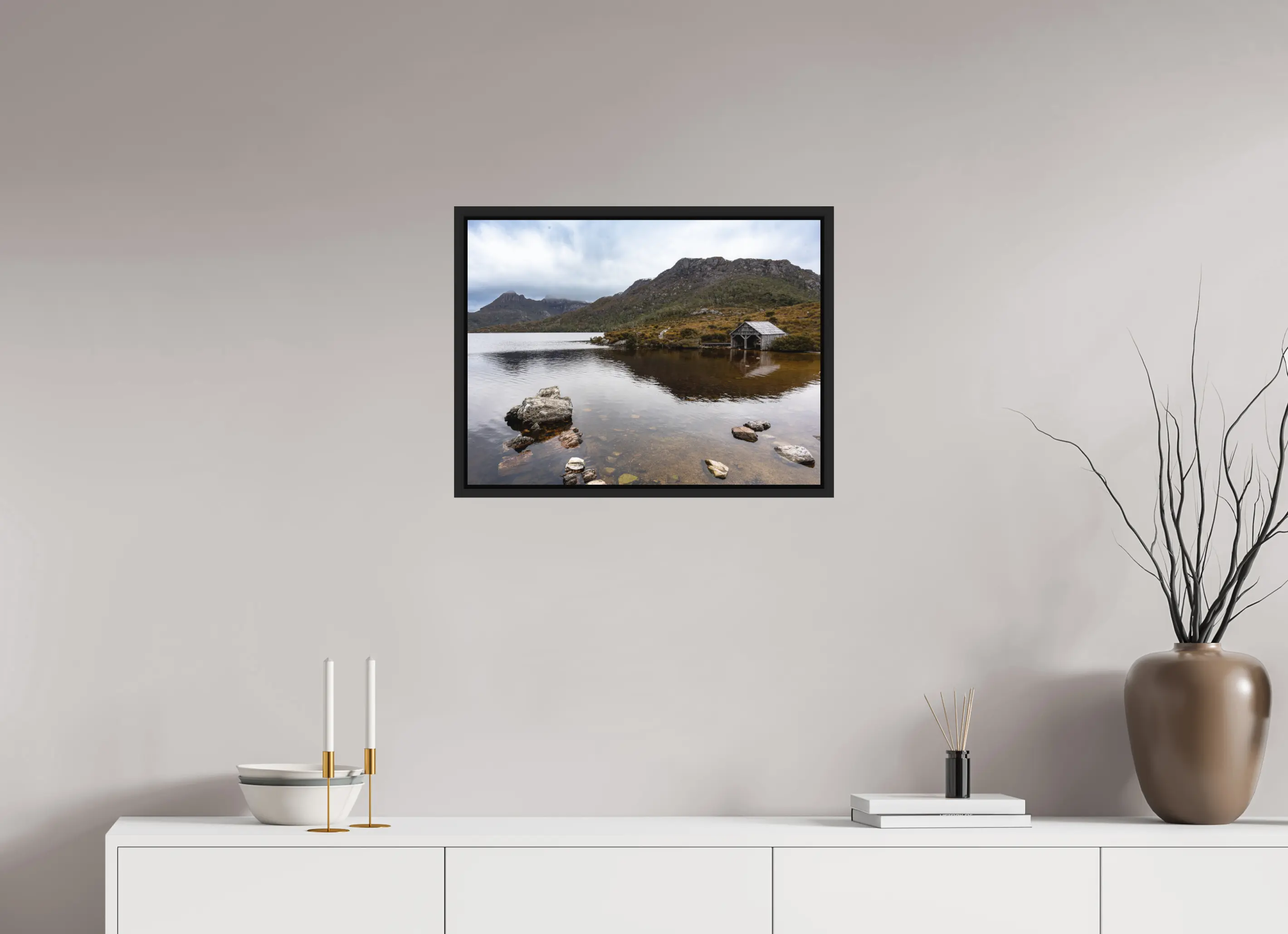 Fine art print “Presence” featuring a wooden boathouse on the calm waters of Dove Lake beneath Cradle Mountain in Tasmania, displayed in a floater frame in a modern living room setting.