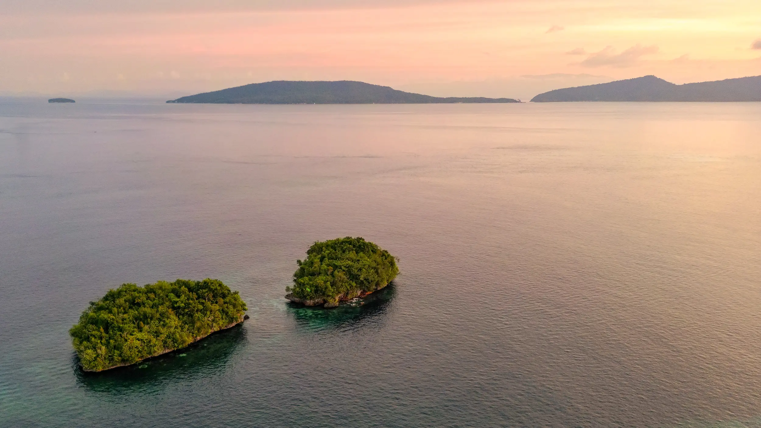 Aerial view of two small lush islands surrounded by calm pink waters at sunset in Raja Ampat Indonesia.