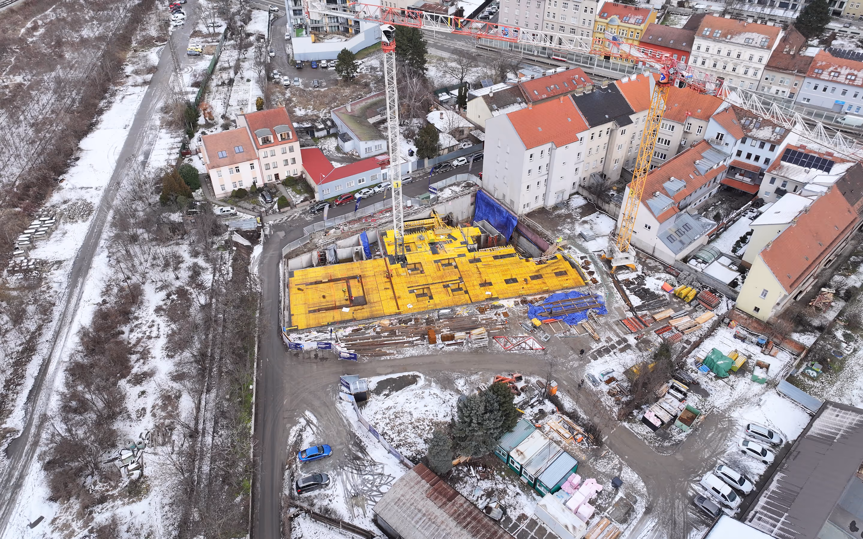 Letecký pohled na stavební místo s žlutou konstrukcí a jeřáby obklopené obytnými domy za zimního počasí.