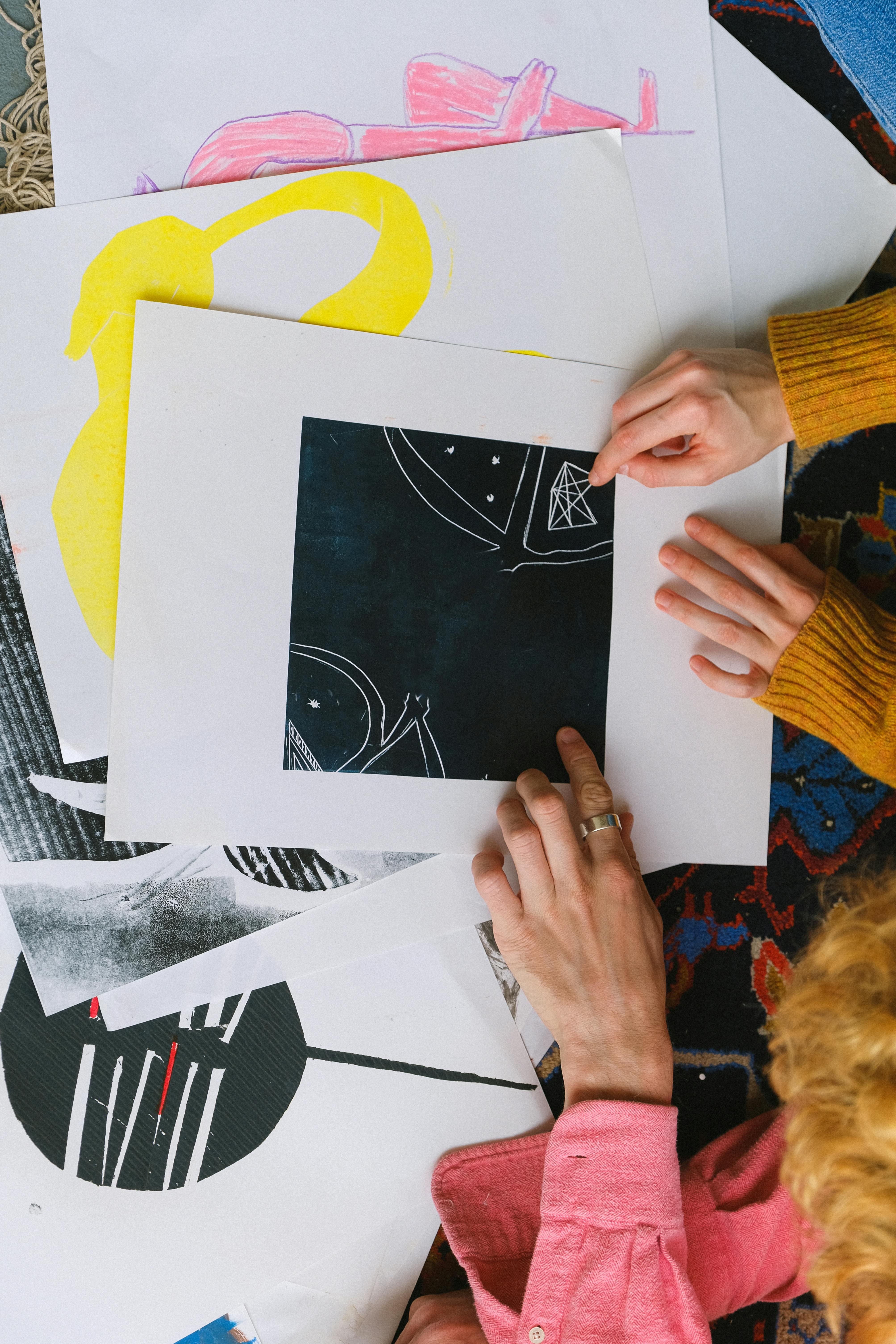 Aerial view of two people pointing at a hand-drawn illustration on a tabletop covered with other illustrations in the same style.