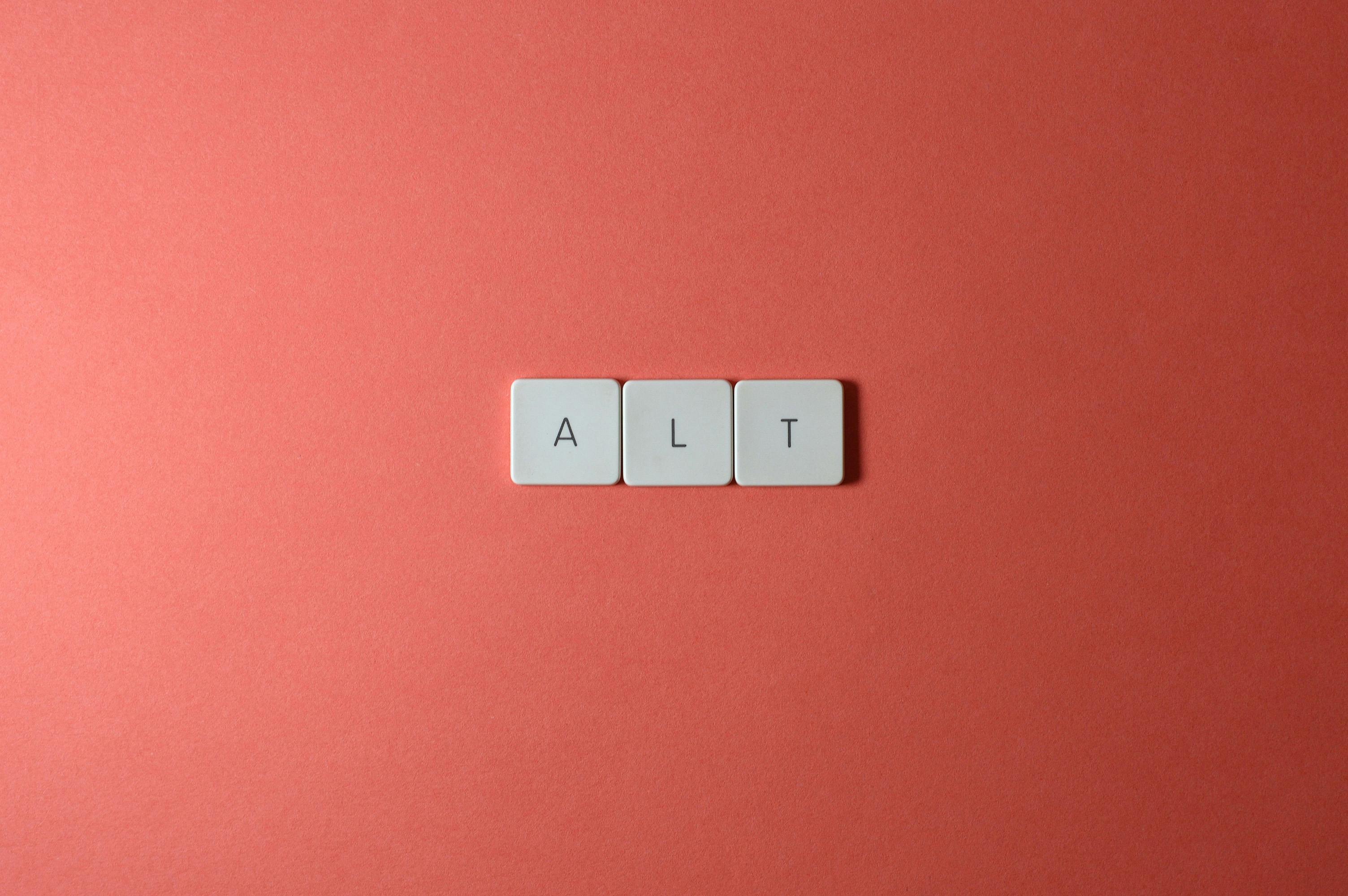 A minimalist photograph shows three white, Scrabble-like tiles that spell the word 'ALT.' The tiles are perfectly centered against a solid coral-colored background.
