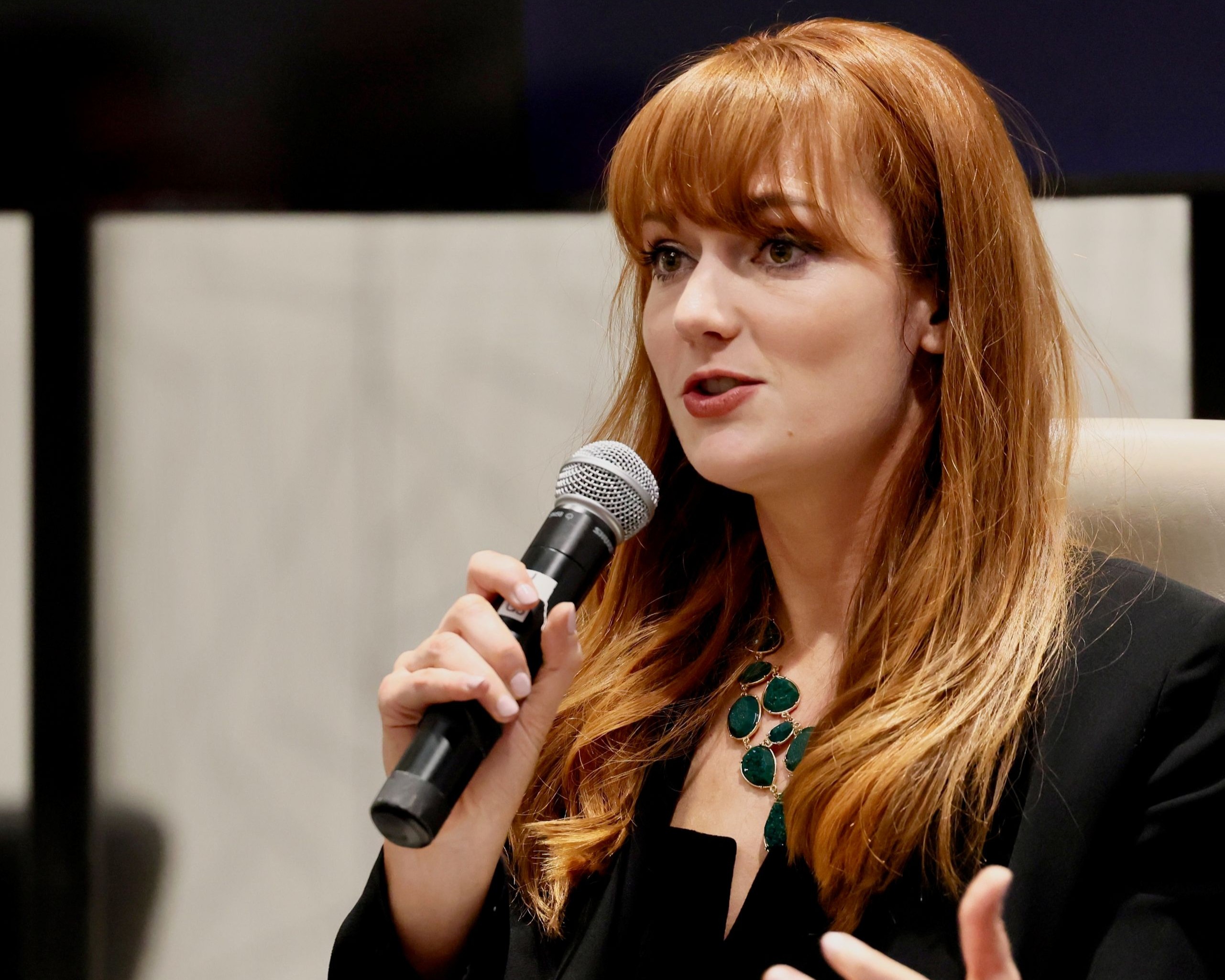 A woman with long, reddish-brown hair and bangs speaks into a microphone. She is wearing a black top and a prominent necklace with large, emerald-green stones. Her right hand holds the microphone, and her left hand is partially visible, gesturing as she speaks. The background is a blurred indoor setting with neutral tones.