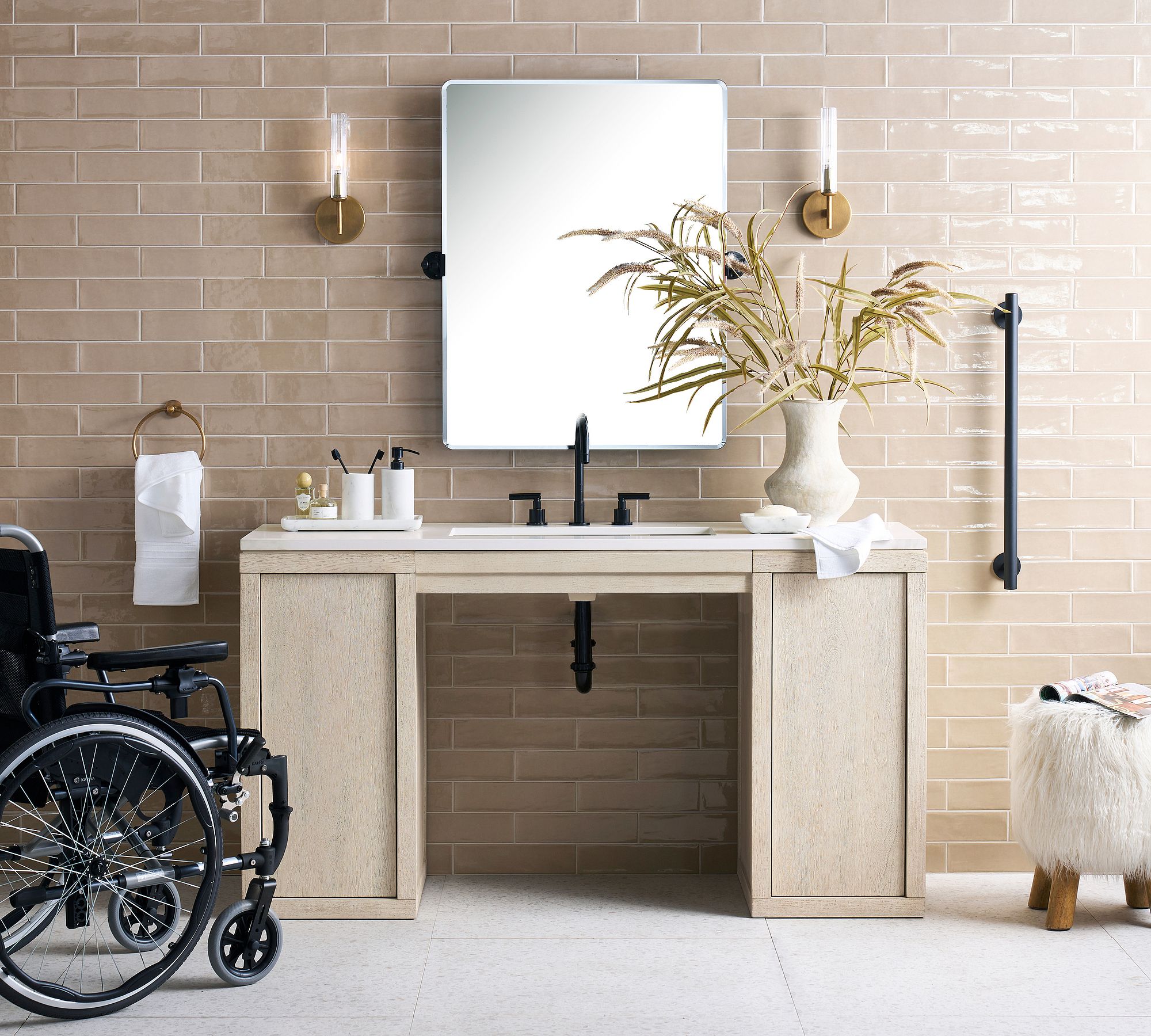 A full bathroom setting featuring the Cayman 61" vanity. The ADA-compliant open-base design is highlighted by a wheelchair positioned to the left. The view is straight-on, showing tan subway tiles, a rectangular mirror, black fixtures, and decorative items.