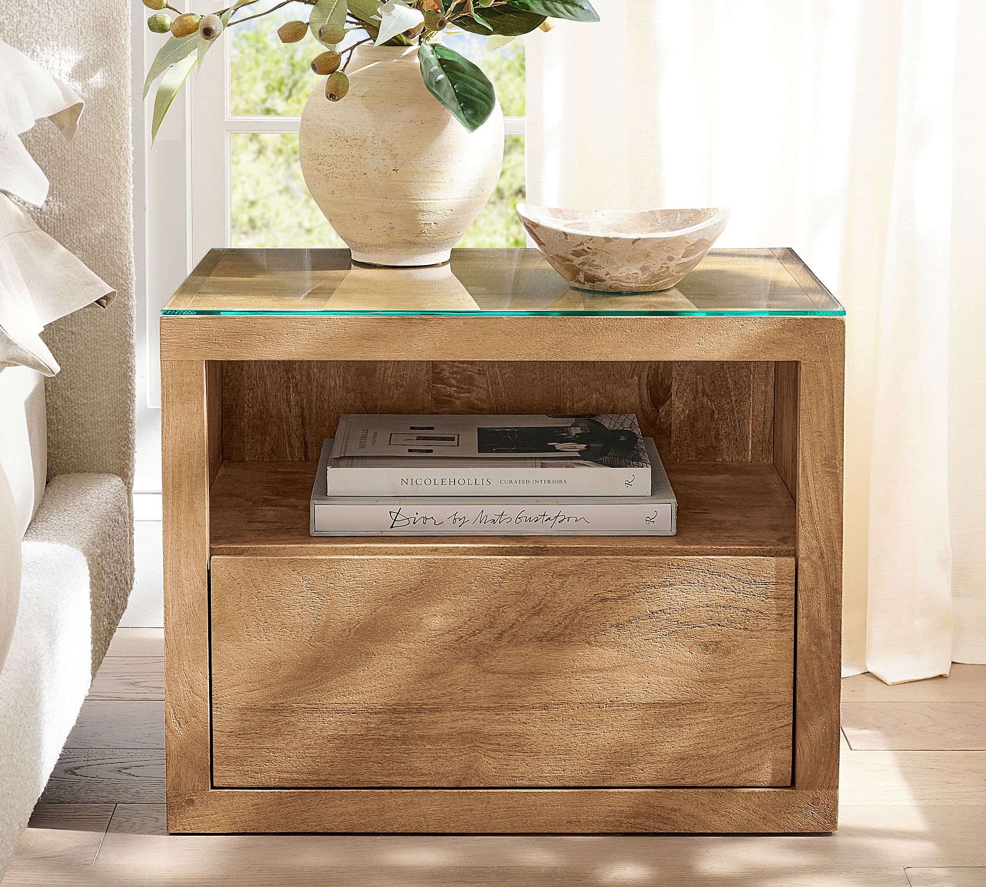 The Cayman Nightstand with a glass top and medium wood finish is positioned next to a bed whose mattress is even with the nightstand's top, illustrating its scale. The nightstand features one open shelf with two stacked books and a single drawer below.