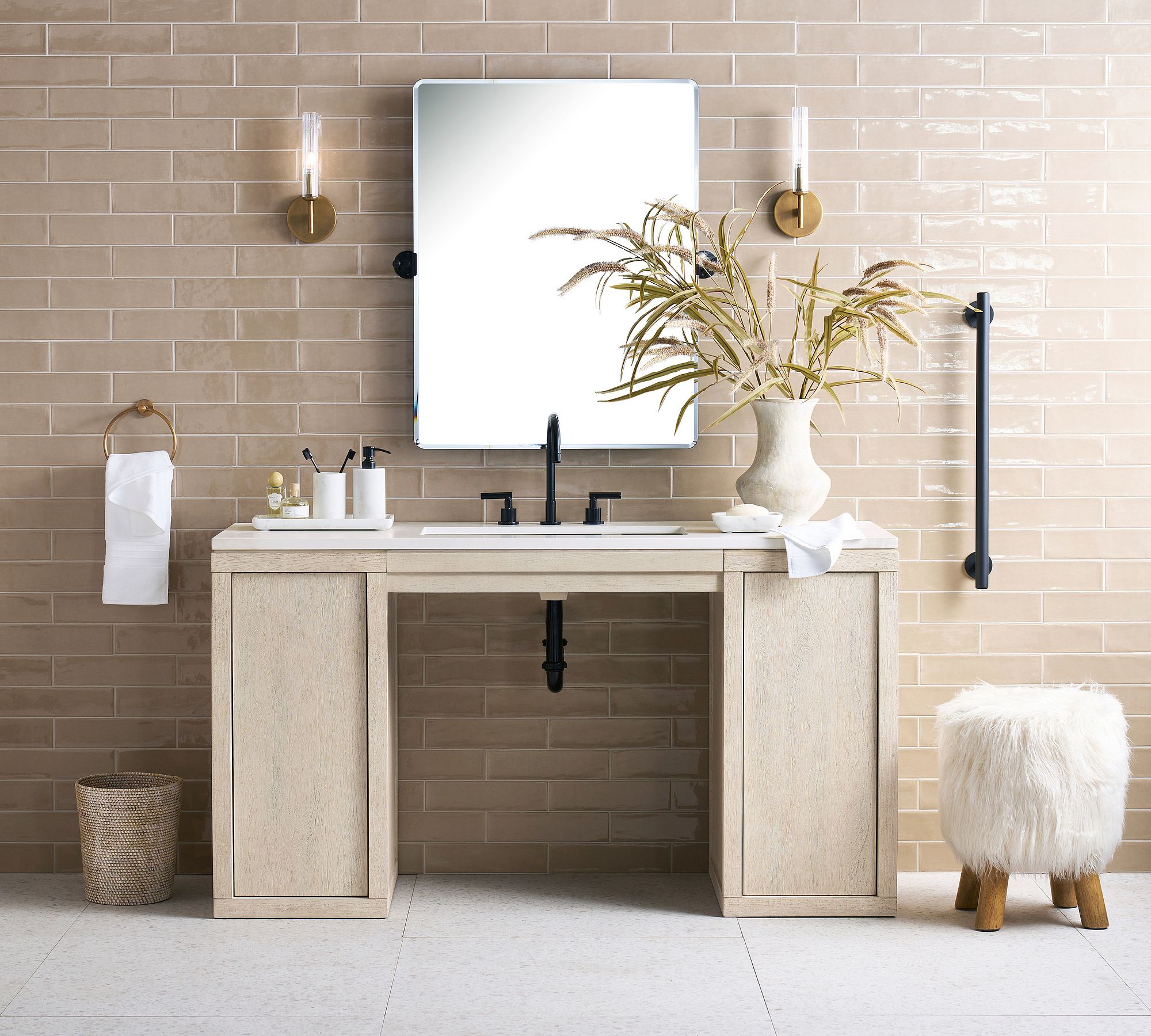 A bathroom setting features the vanity, styled beneath the Vintage Rounded Rectangular Pivot Mirror and Cooper Ribbed Sconces. The background features glossy, tan subway tiles. The Mongolian Faux Fur Stool and Linden Grab Bar are to the right.