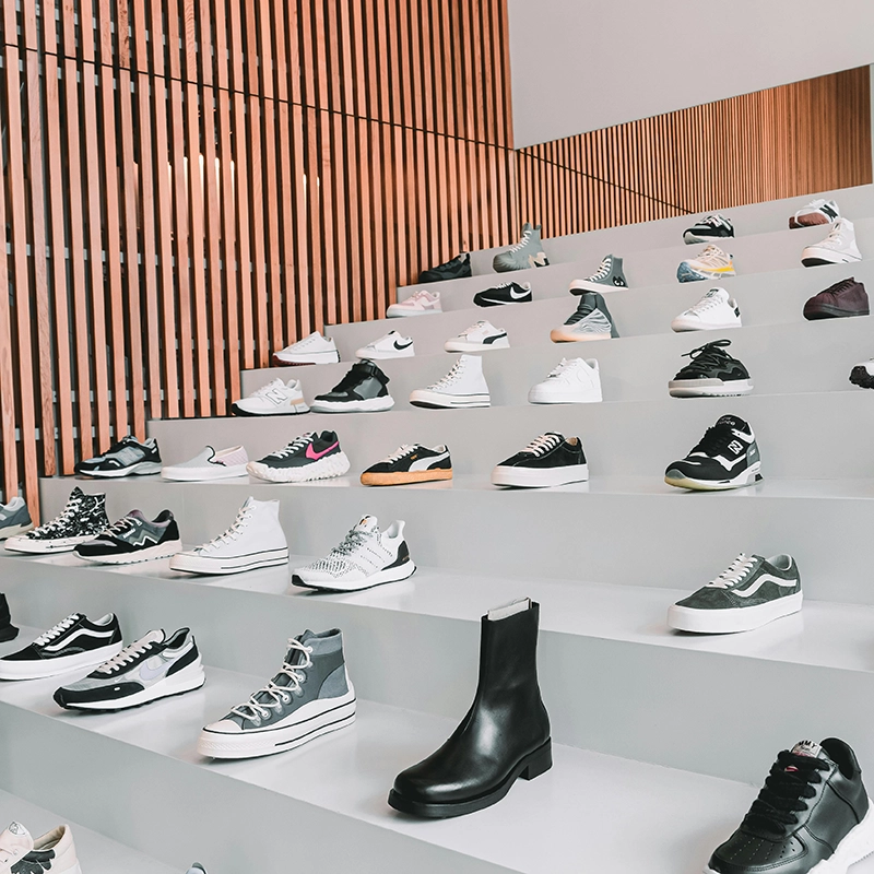 A sleek retail display featuring rows of trendy shoes on a multi-tiered white platform, set against a modern backdrop of high ceilings and vertical wood paneling.
