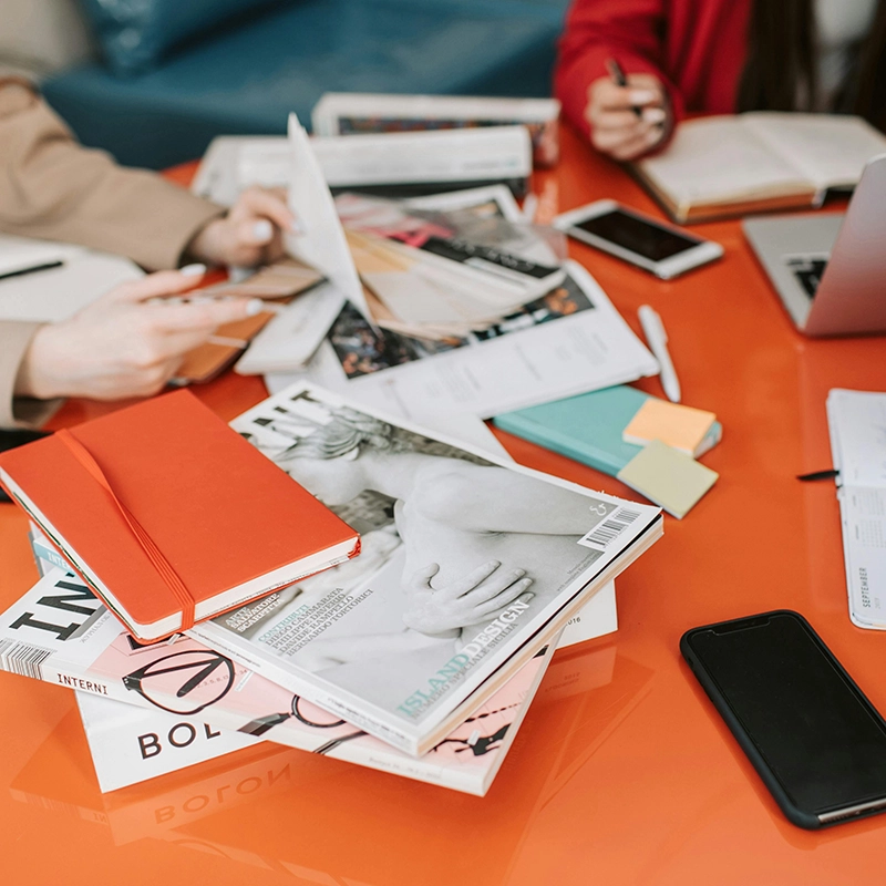 Three creatives at work collaborating around a bright orange desktop covered in photographic prints or graphic designs, utilizing laptops, smartphones, and notepads.