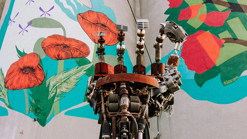 A mechanical humanoid robot hand, with visible internal gears, reaching upward against a vibrant, hand-painted background of colorful poppies and green ferns.