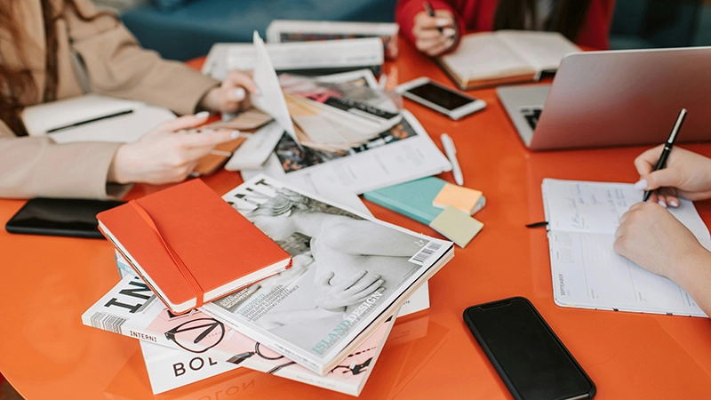 Three creatives at work collaborating around a bright orange desktop covered in photographic prints or graphic designs, utilizing laptops, smartphones, and notepads.
