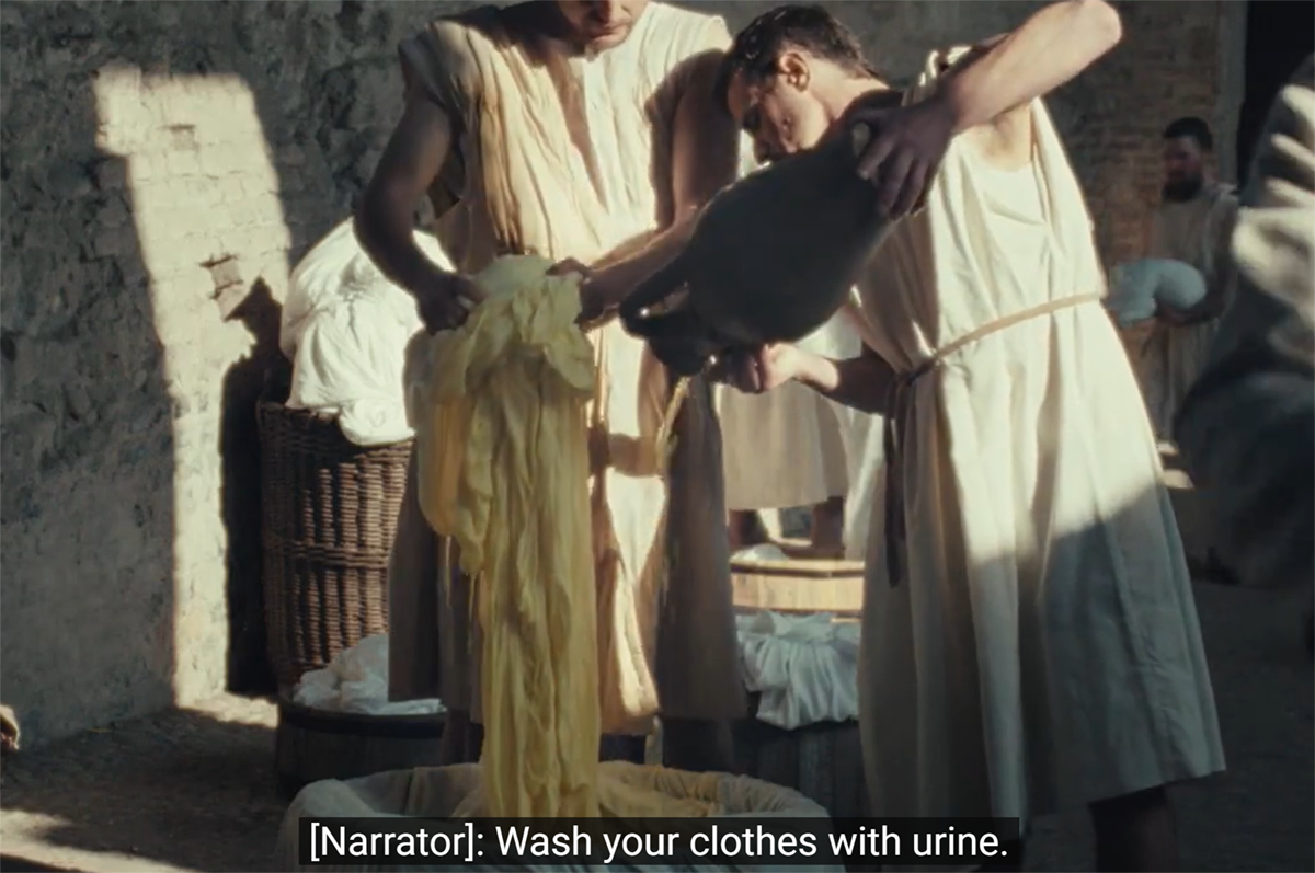Campaign still frame shows two men in togas, one pouring liquid from a jug, the other holding yellow sheets. Caption reads, "[Narrator]: Wash your clothes with urine."