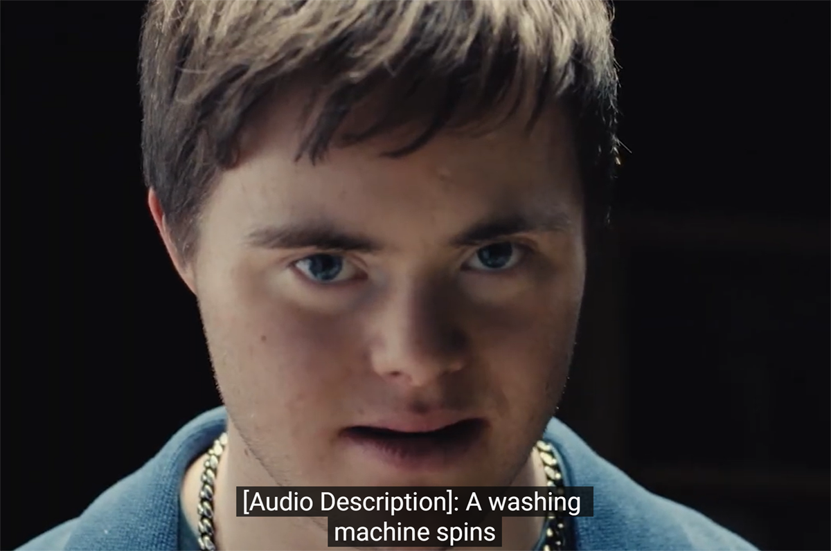Campaign still frame shows a close-up of our narrator looking directly at us with a serious expression. Caption reads, "[Audio Description]: A washing mashine spins..."