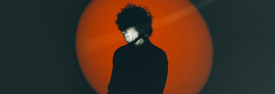 Silhouette of a Enrique Bunbury standing against a large, glowing orange circle on a dark background.