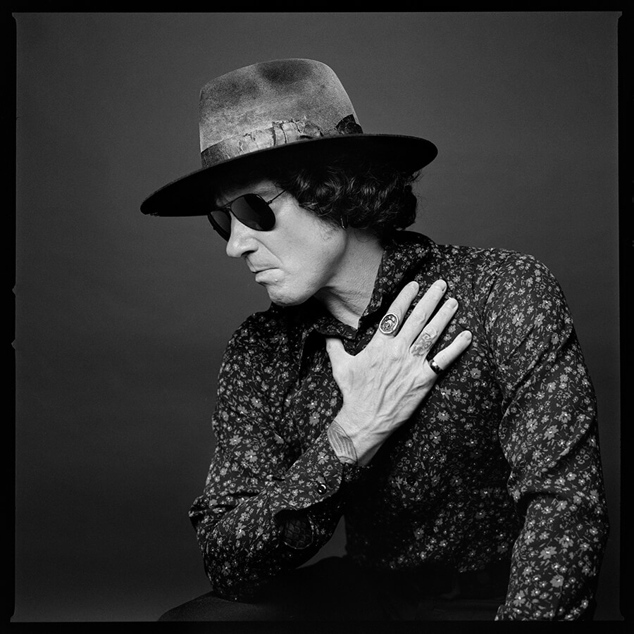 Black and white portrait of a Enrique Bunbury in a wide-brimmed hat, sunglasses, and floral shirt with his hand on his chest.
