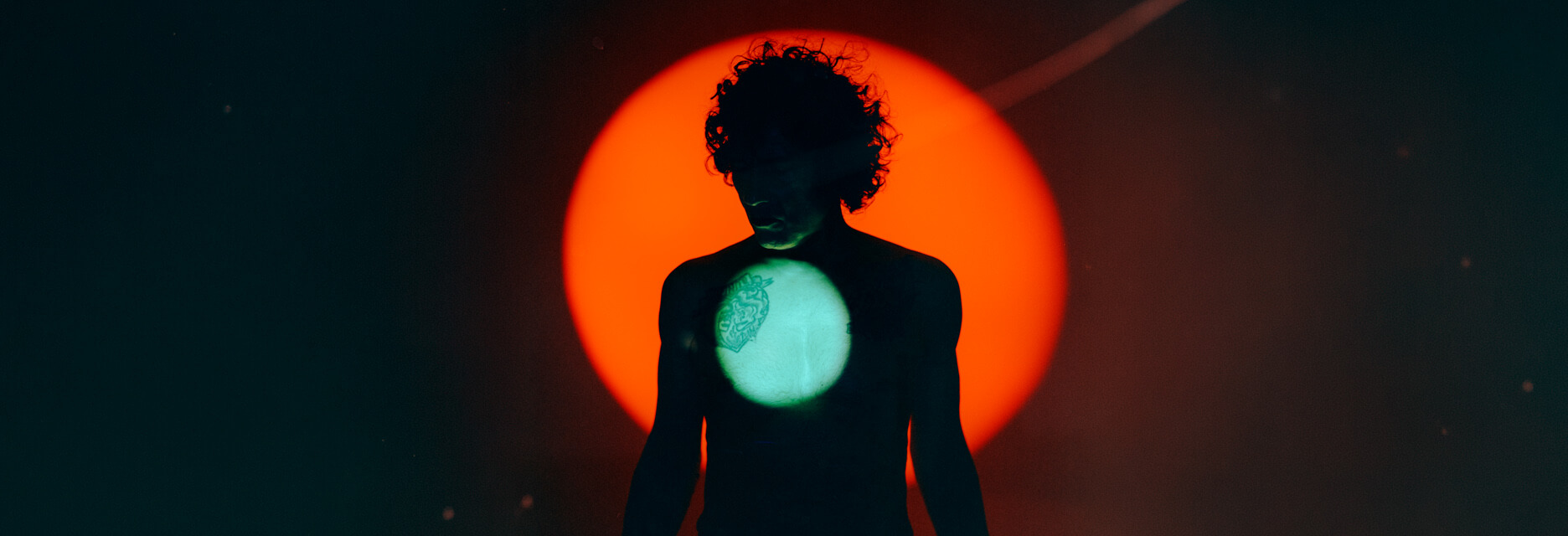 Silhouette of Enrique Bunbury with a tattoo on their chest, lit by a bright red circular light behind and a smaller greenish light on their chest.