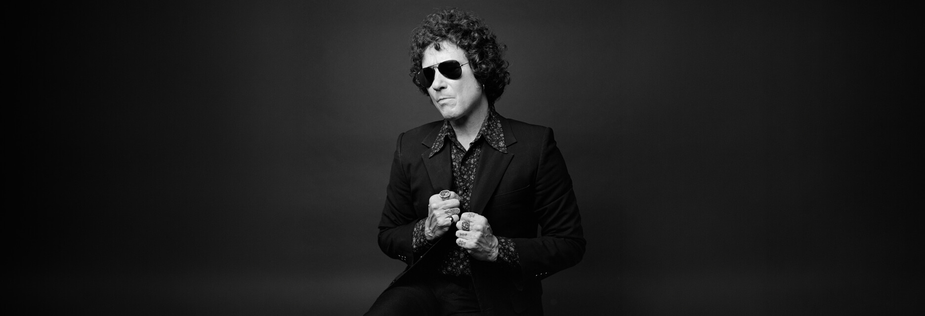 Black and white portrait of Enrique Bunbury wearing sunglasses and a dark suit, clasping his hands in front of him.