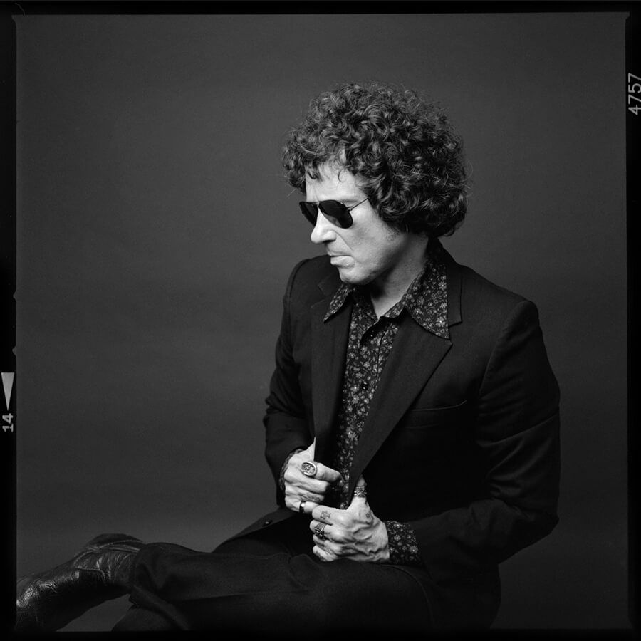 Black and white portrait of Enrique Bunbury wearing sunglasses, a patterned shirt, and a blazer, sitting with legs crossed.