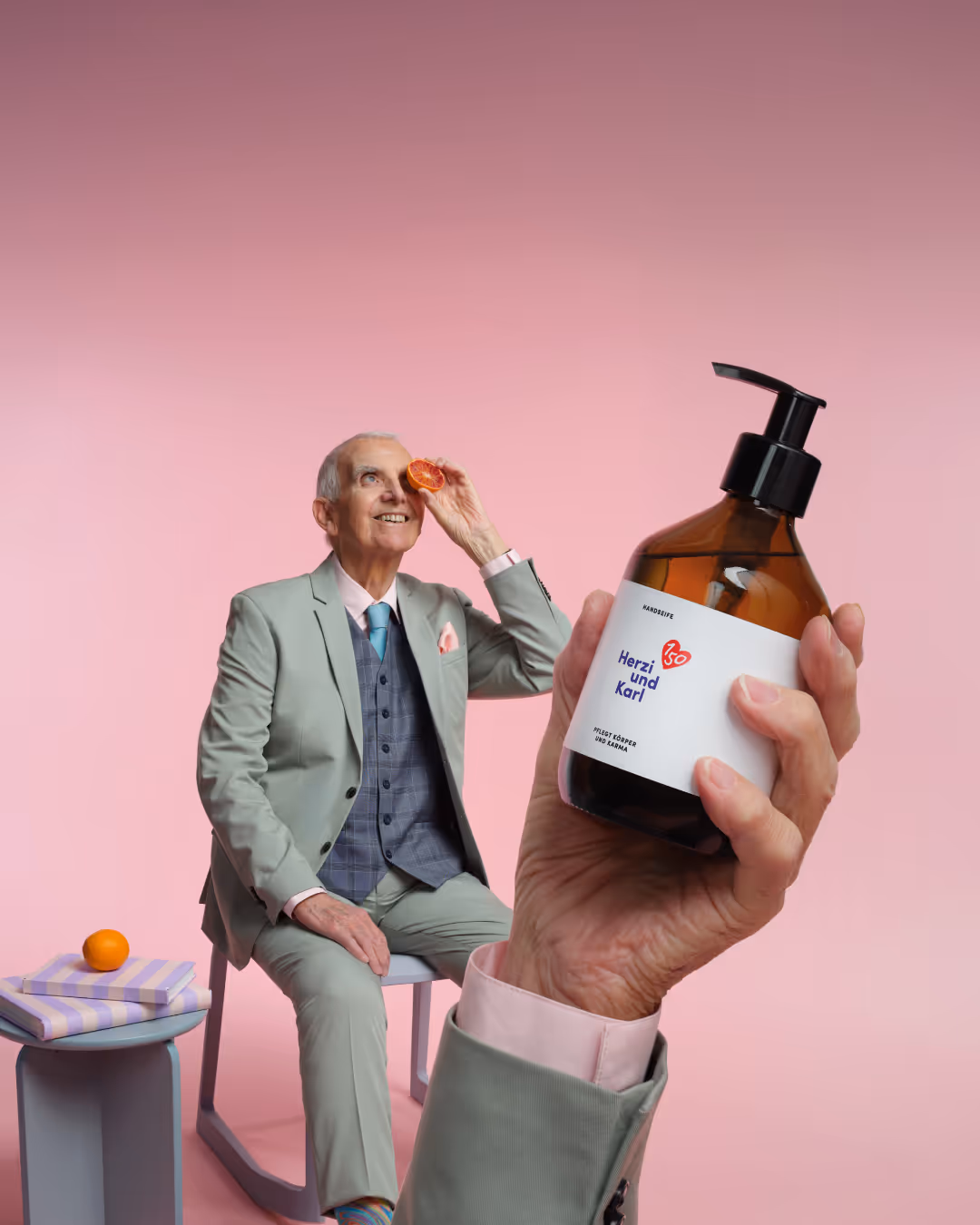 Elderly gentleman in pastel green suit holds a sliced blood orange to his eye, while a hand in the foreground presents the anniversary hand soap "Herzi and Karl" from Haus der Barmherzigkeit x Saint Charles. Friendly pink background emphasizing the celebration of the 150th anniversary of House of Mercy. Anniversary campaign of the House of Mercy, realized by the lead agency Achtzehn Grad from Vienna.