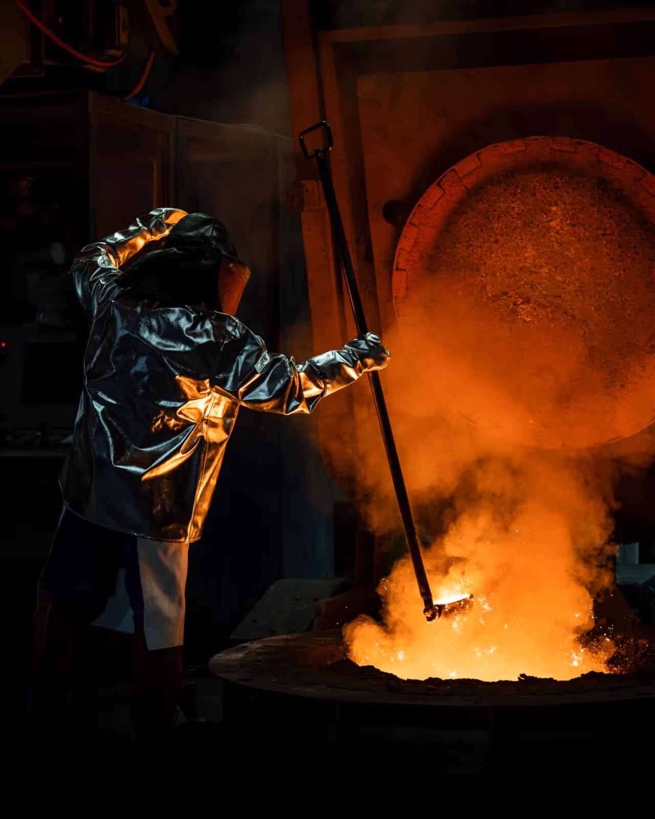 FRISCHHUT-Mitarbeiter in hitzebeständiger Schutzkleidung überwacht den Gussvorgang an einem Schmelzofen – glühendes Eisen, Funkenflug und präzise Handarbeit in der Produktion hochwertiger Kanalgussformstücke.