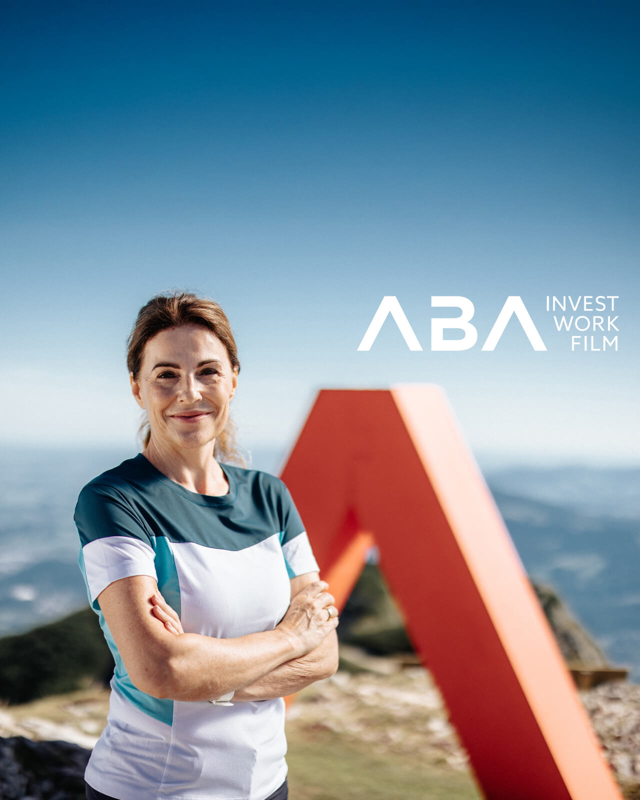 Eine sportlich gekleidete Frau steht mit verschränkten Armen lächelnd im Vordergrund, im Hintergrund ist ein großes rotes „A“-Sculpture, stellvertretend für die Austrian Business Agency (ABA), auf einem Berggipfel zu sehen. Rechts oben im Bild befindet sich das Logo „ABA INVEST WORK FILM“ vor einem klaren blauen Himmel.