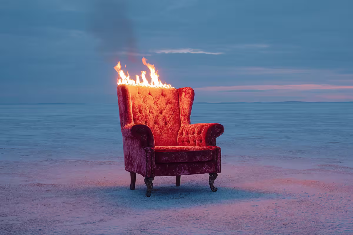 Roter Vintage-Sessel steht allein in einer weiten, blauen Landschaft, während seine Rückenlehne hell brennt – surrealistische Szene mit starkem Farbkontrast